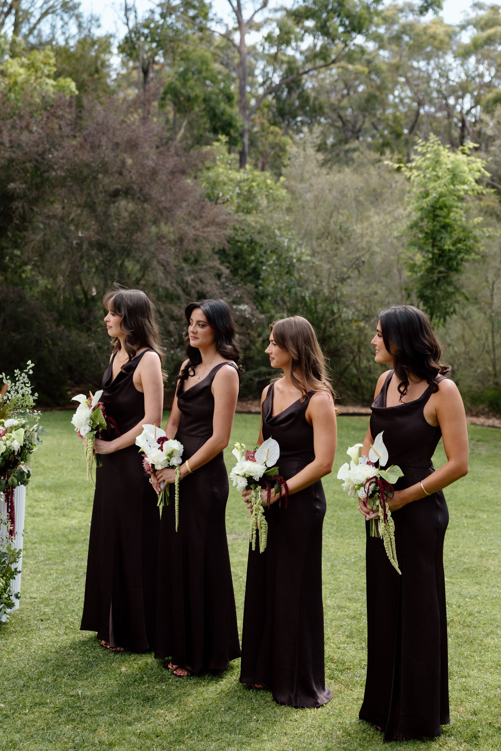 Four bridesmaids in dark dresses holding bouquets stand outdoors on grass.