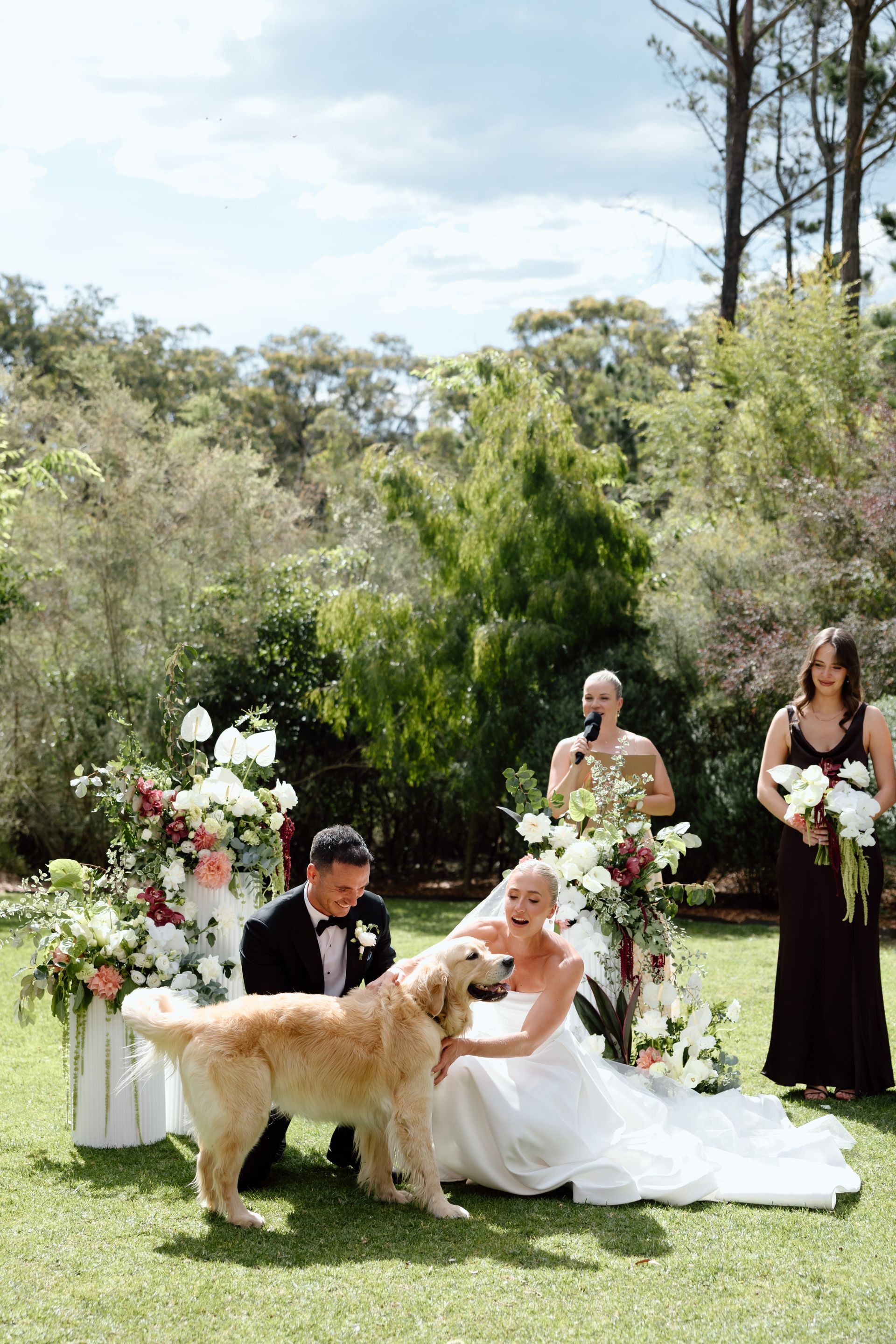 Bride and groom with a golden retriever at an outdoor wedding, surrounded by floral arrangements.