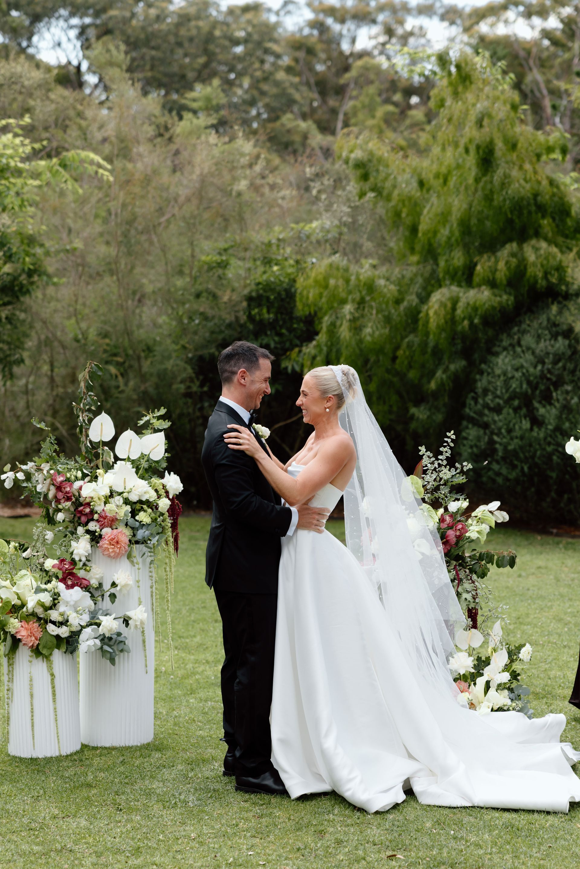 Bride and groom embrace during an outdoor wedding ceremony, with floral arrangements and greenery around them.