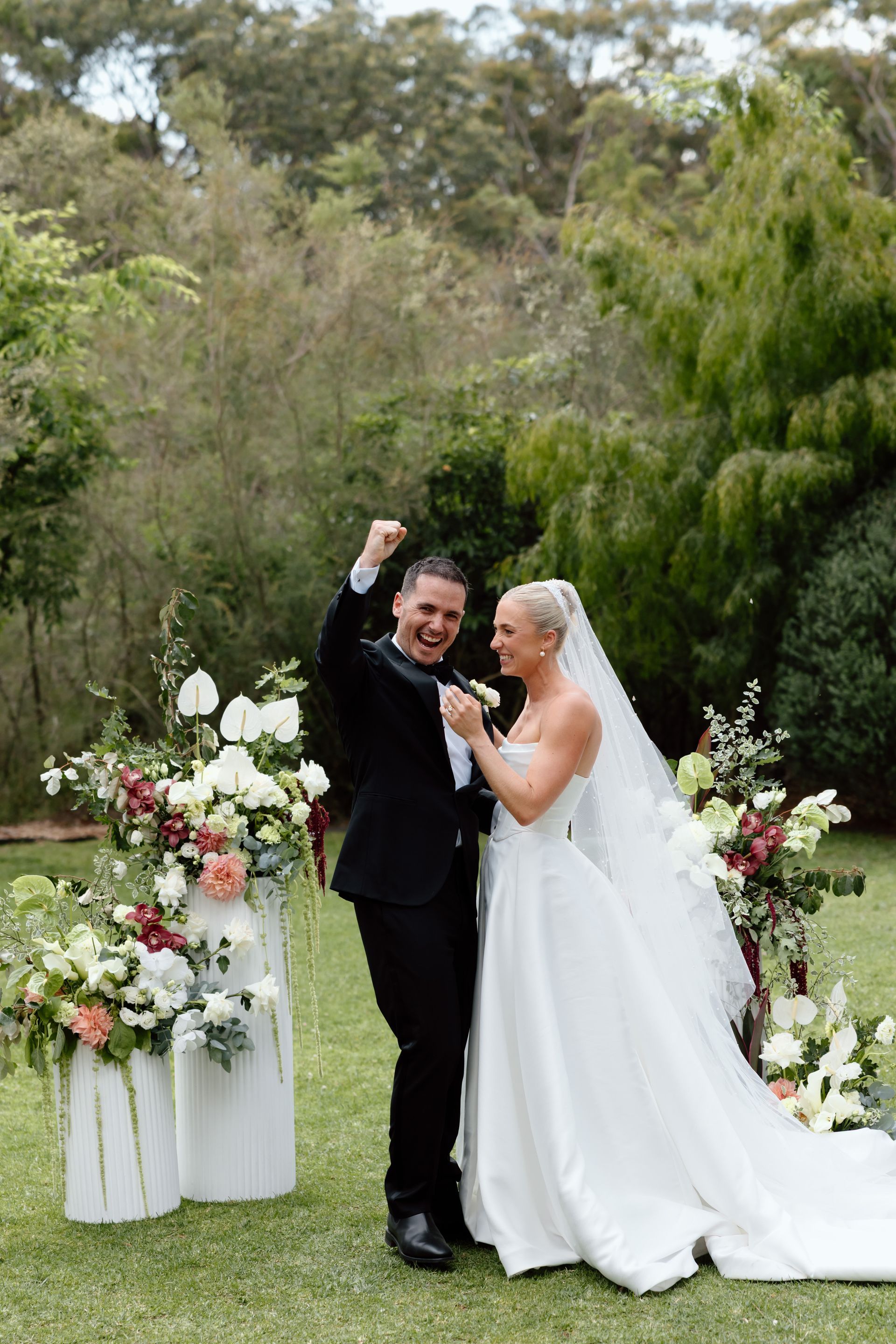 Bride and groom posing outdoors at a wedding ceremony, smiling beside white floral arrangements.