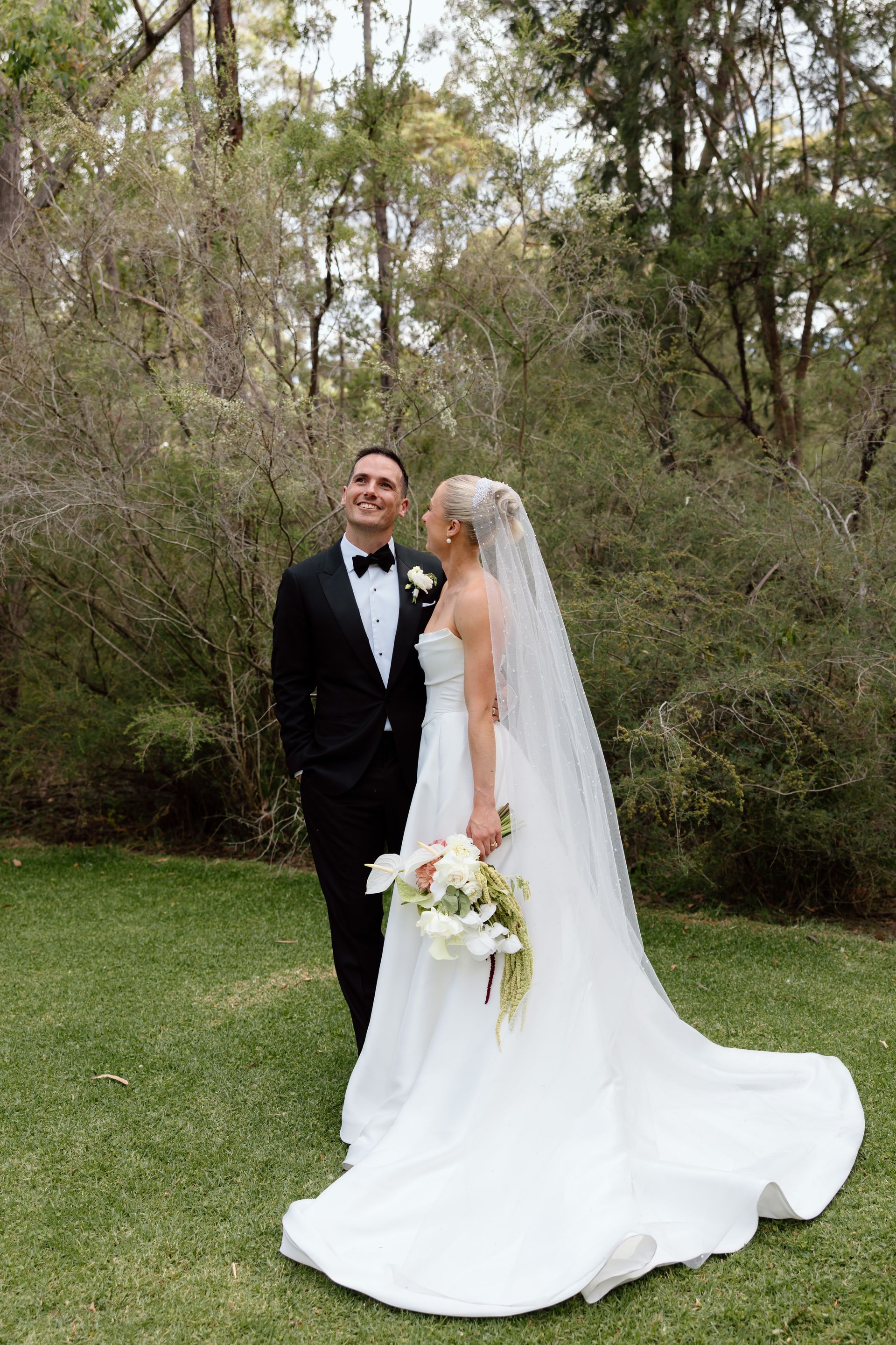Bride and groom posing outdoors in a garden, bride in white gown with long veil and bouquet.