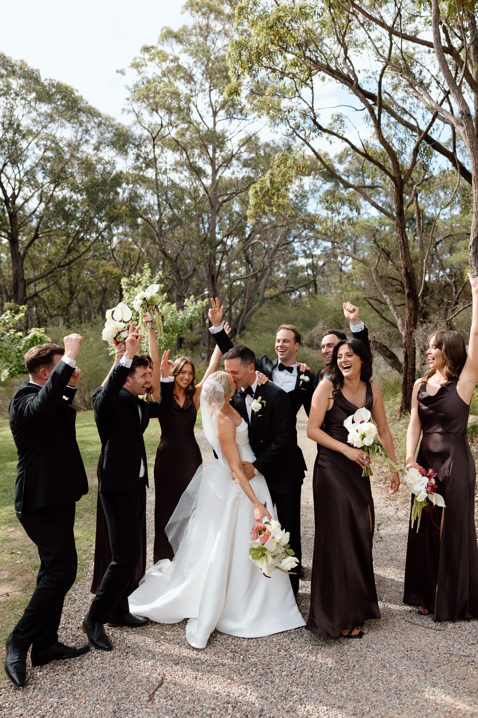 Wedding party celebrating outdoors in a wooded setting, bride in white surrounded by attendants in black gowns and suits