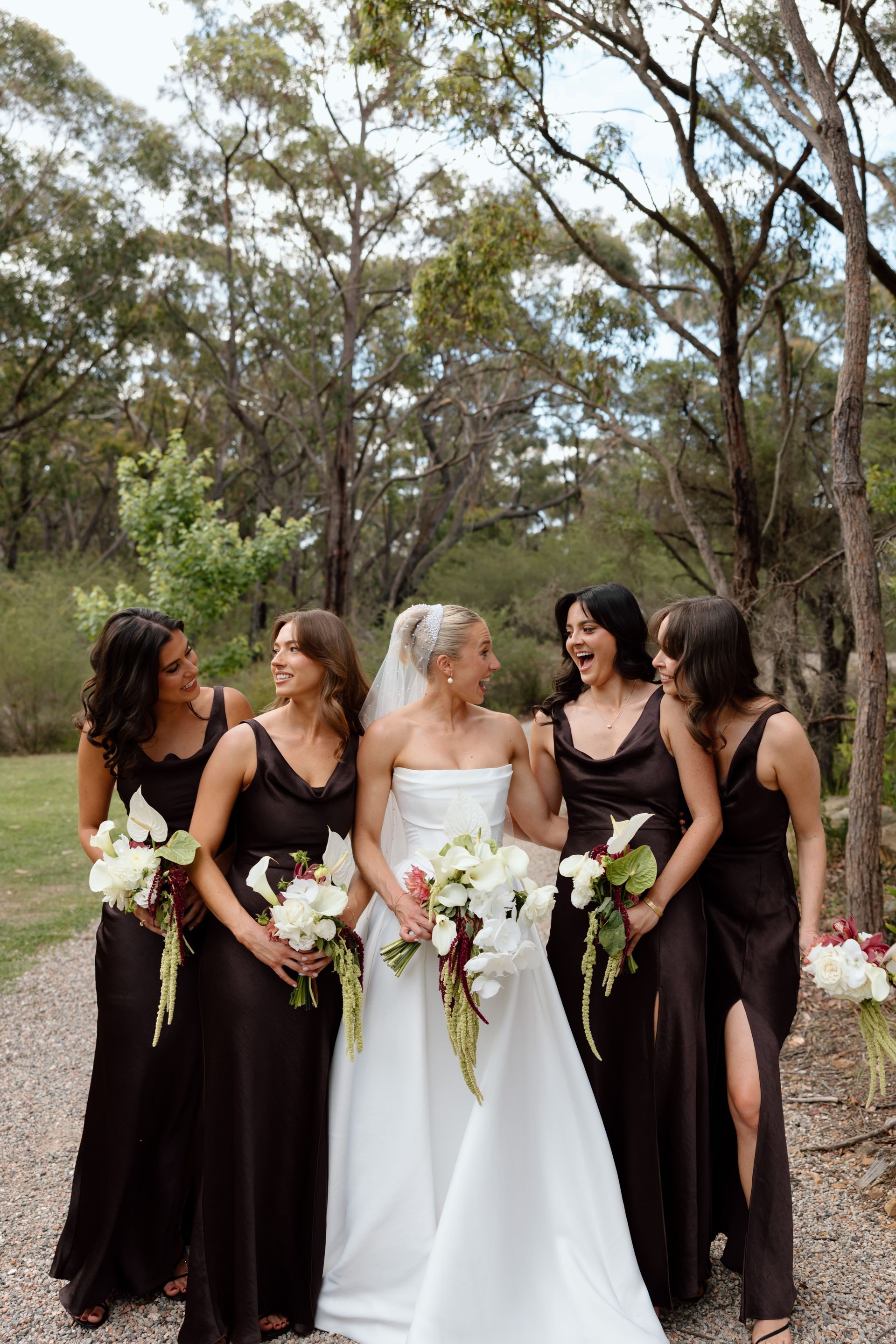 Five women in black and white dresses holding bouquets outdoors in a wooded setting