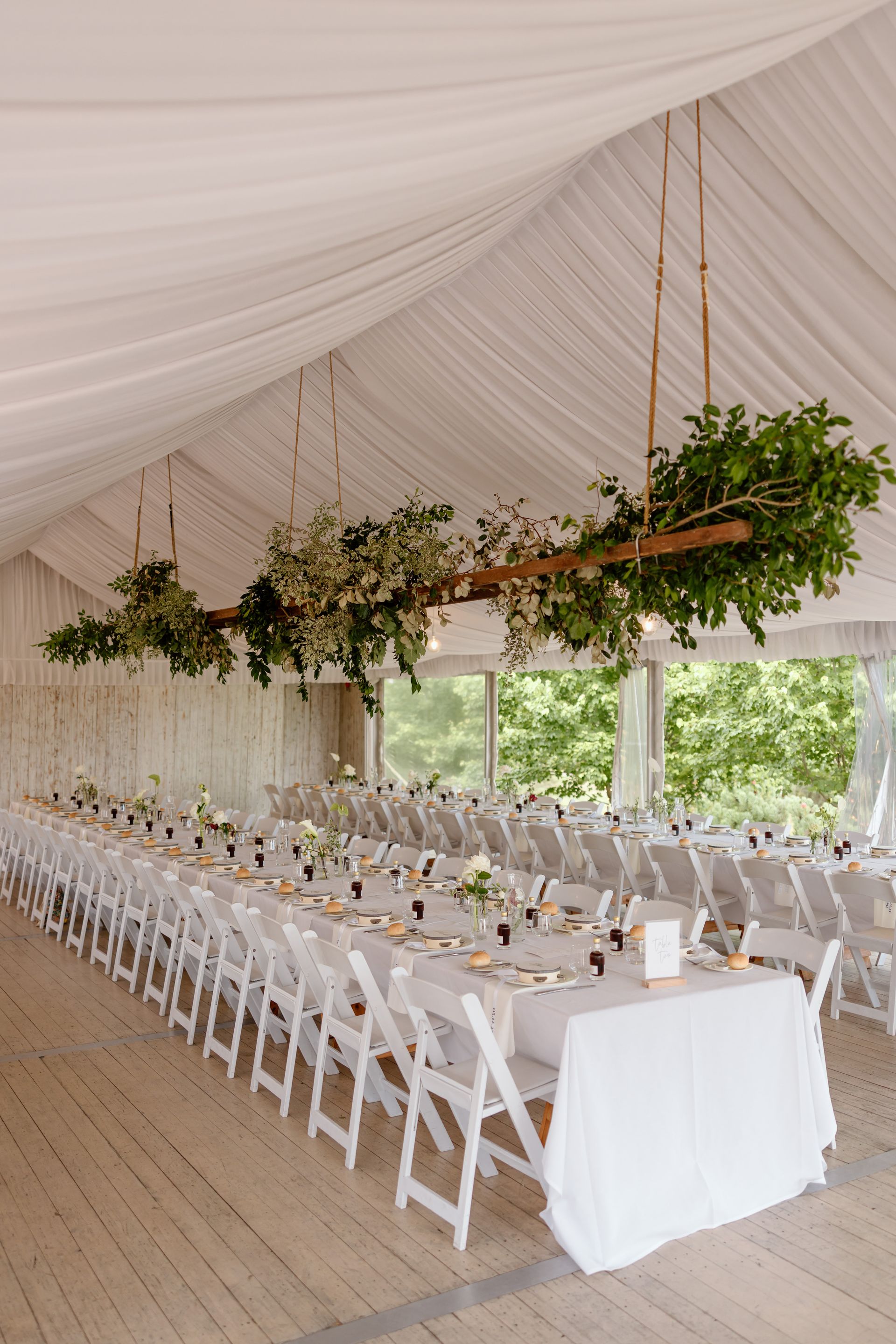 Elegant tented reception with long white table, folding chairs, and hanging greenery decorations