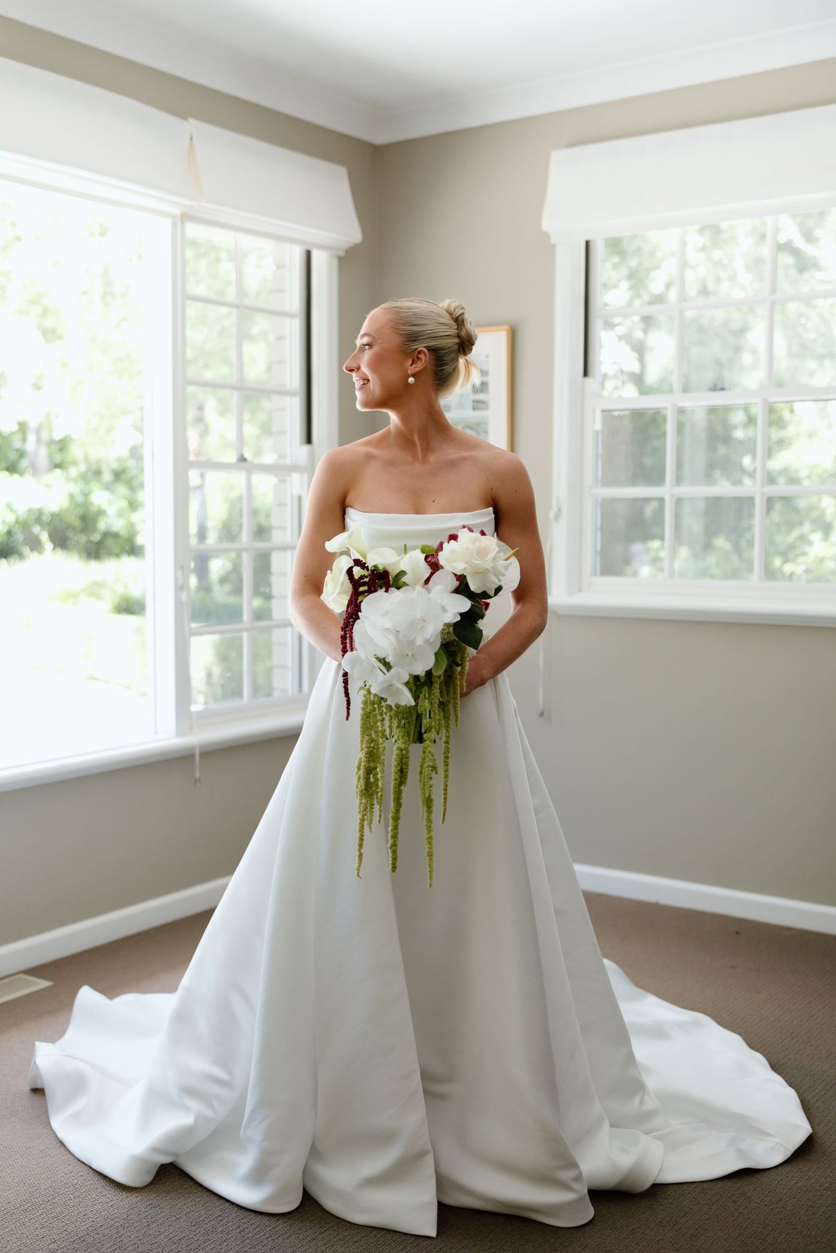 Bride in a white gown holding a orchid bouquet by bright windows indoors