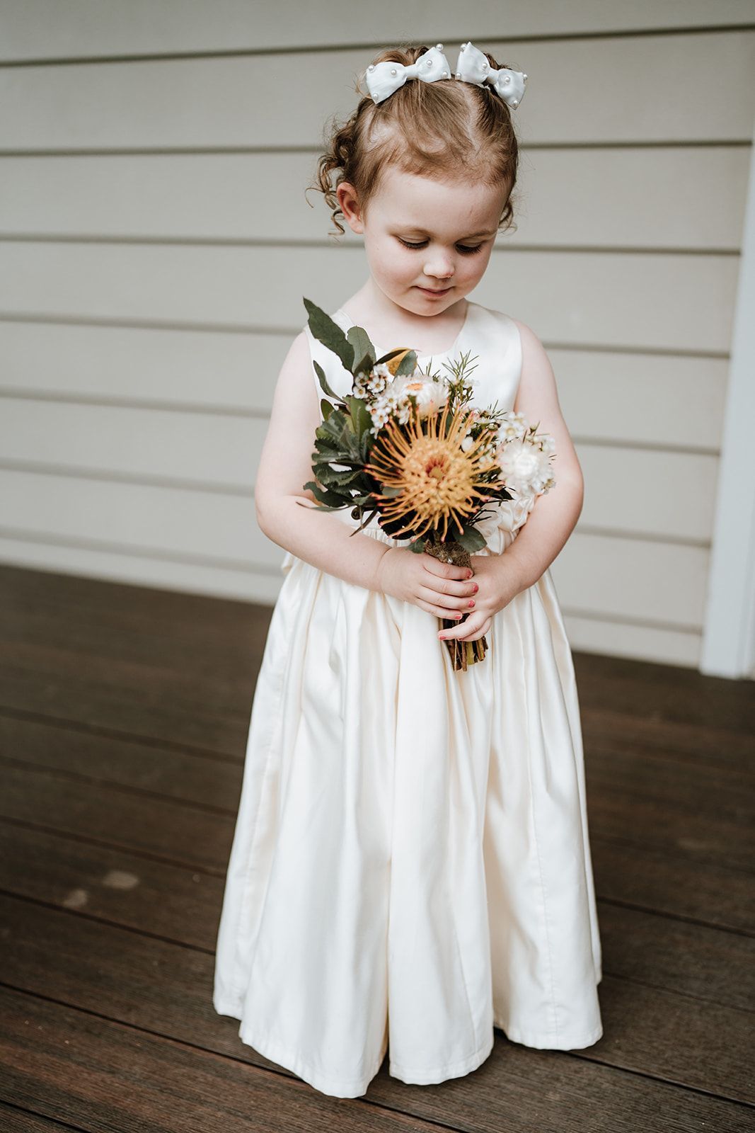 Young child in a white dress holding a bouquet of flowers on a porch, looking down.