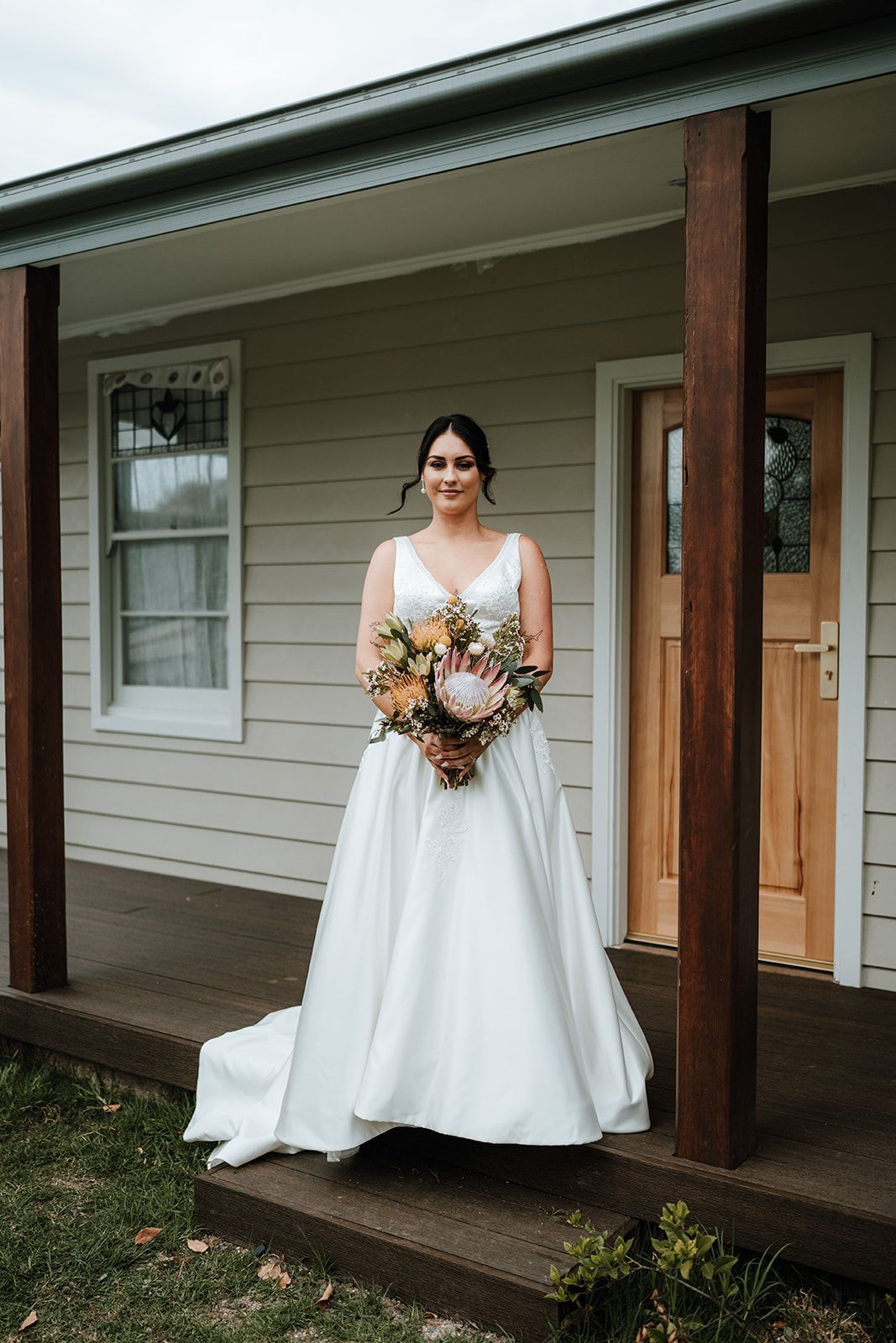 Bride in a white gown holding a bouquet, standing on a porch in front of a house
