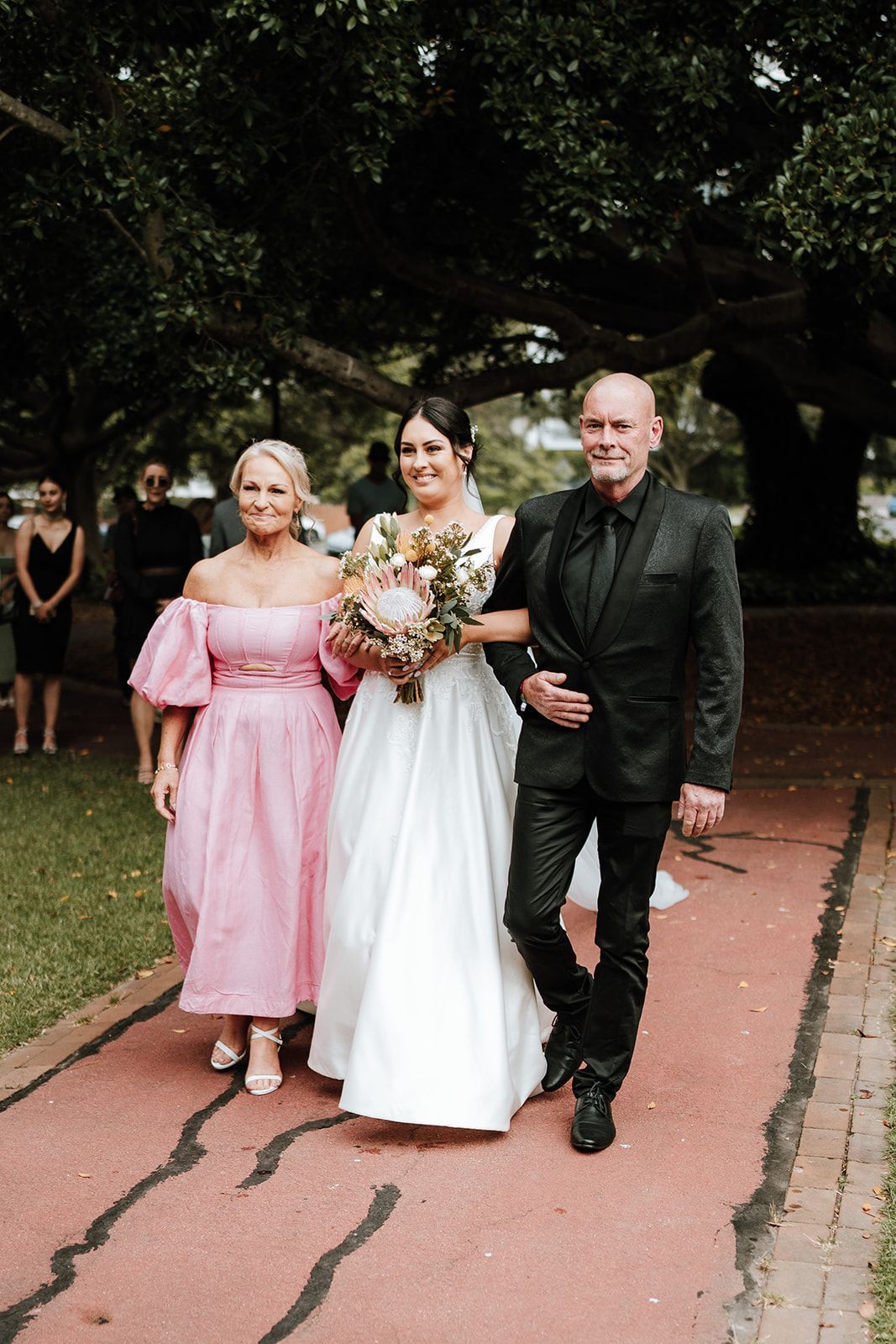 Bride walks with two companions down a garden path, holding a bouquet and wearing a white gown.
