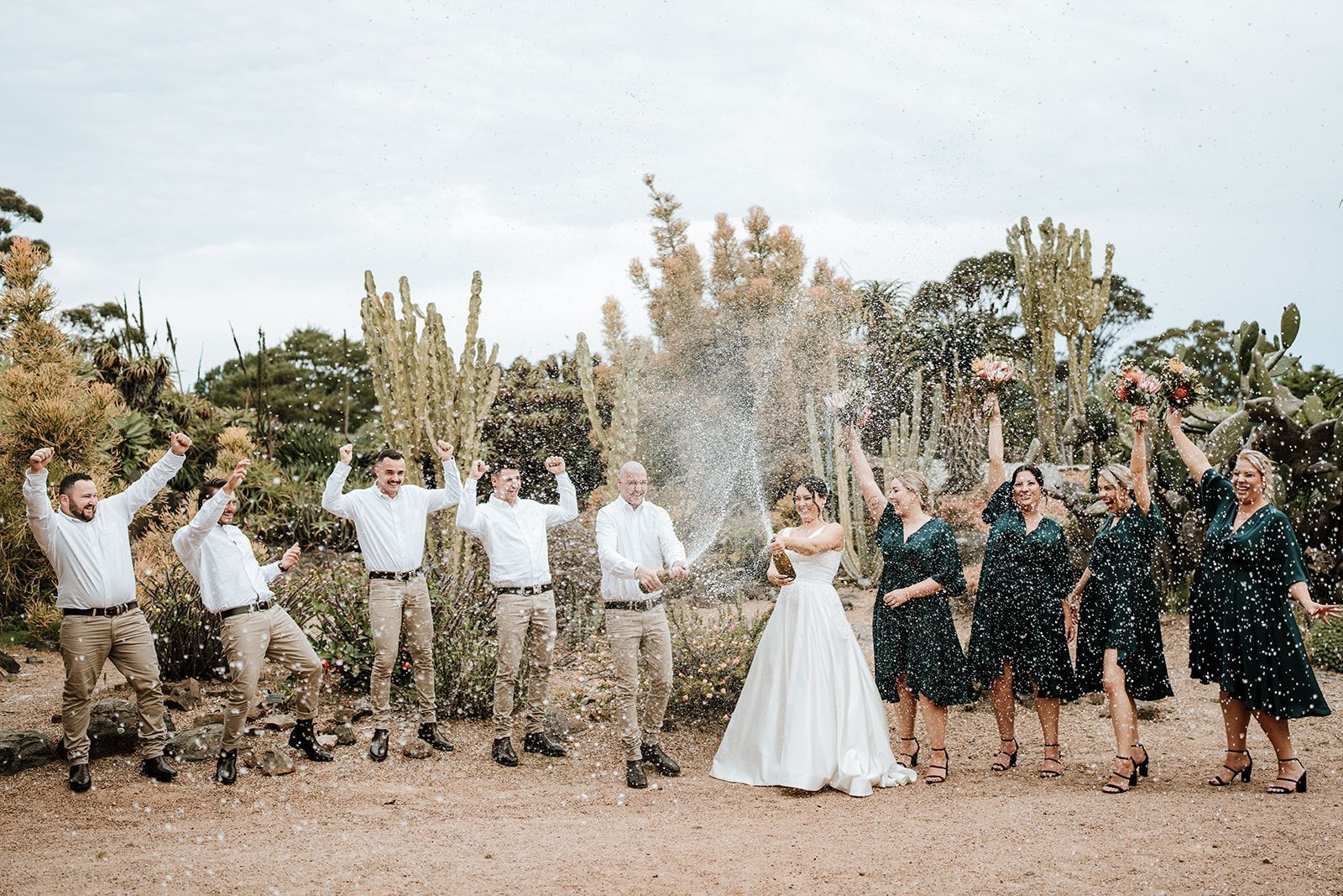 Wedding party celebrating outdoors with champagne spray, bride in white center, guests in dark and light outfits