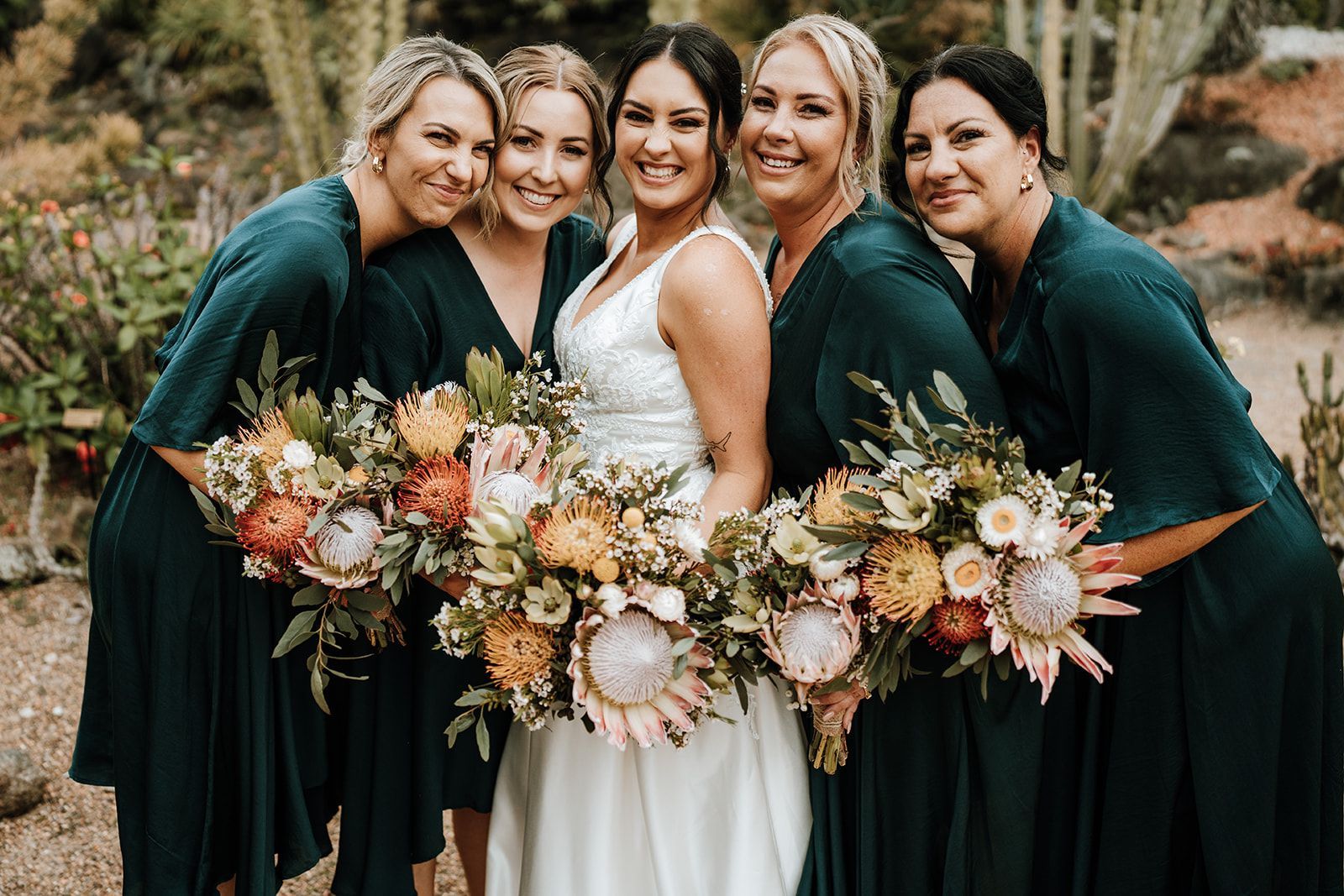 Five people in dark green dresses and a white dress holding autumn bouquets outdoors