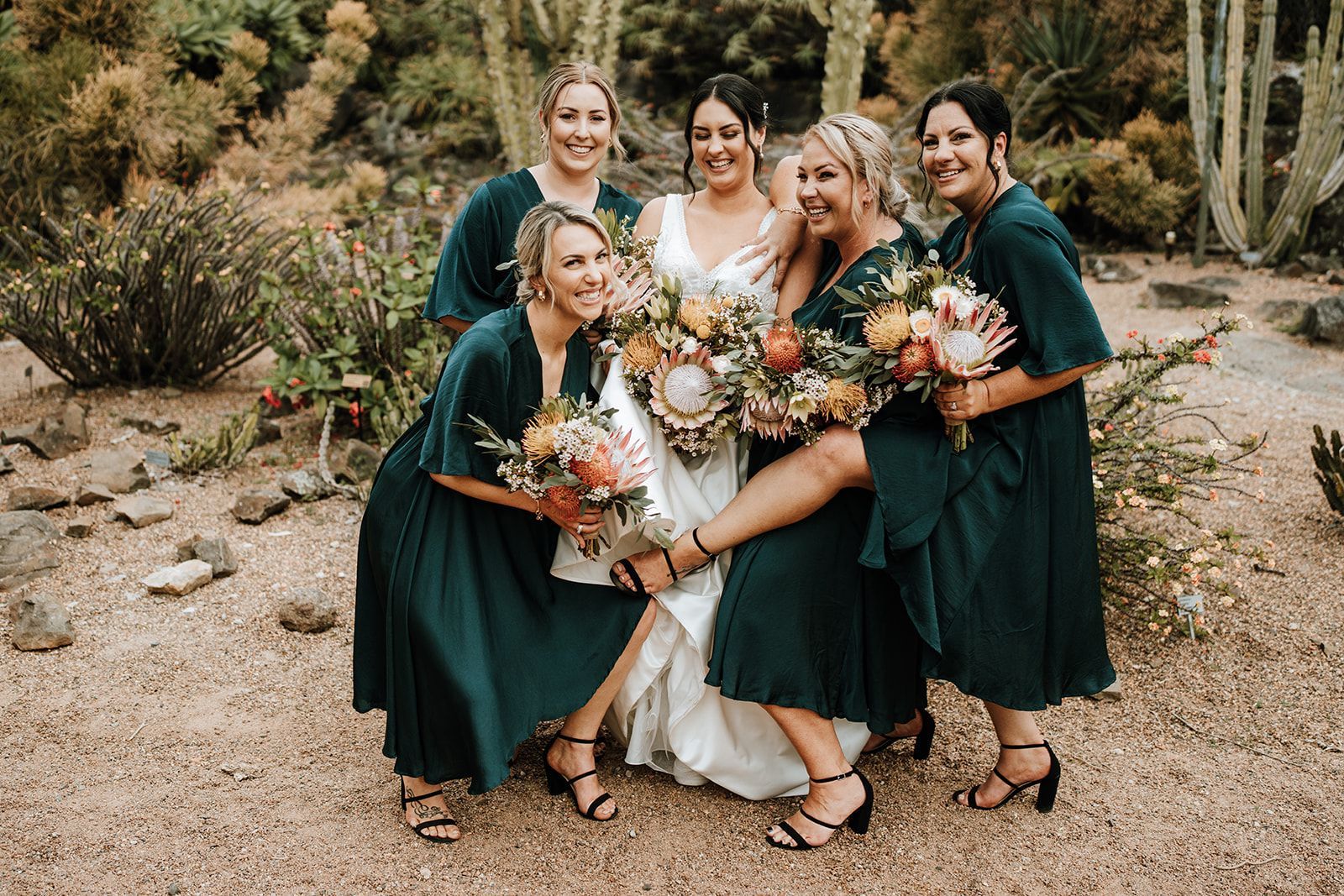 Five people in dark green dresses and a white dress pose outdoors holding bouquets.