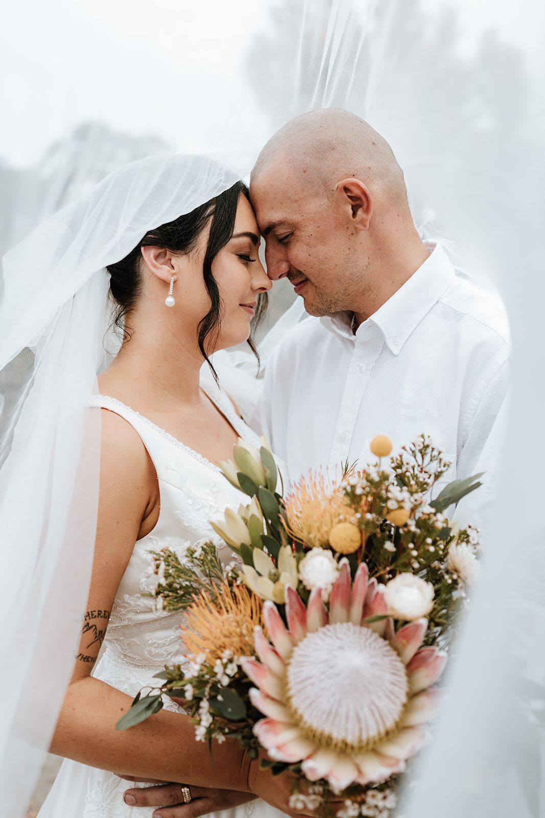 Bride and groom touch foreheads in a soft wedding portrait, holding a pastel bouquet outdoors.