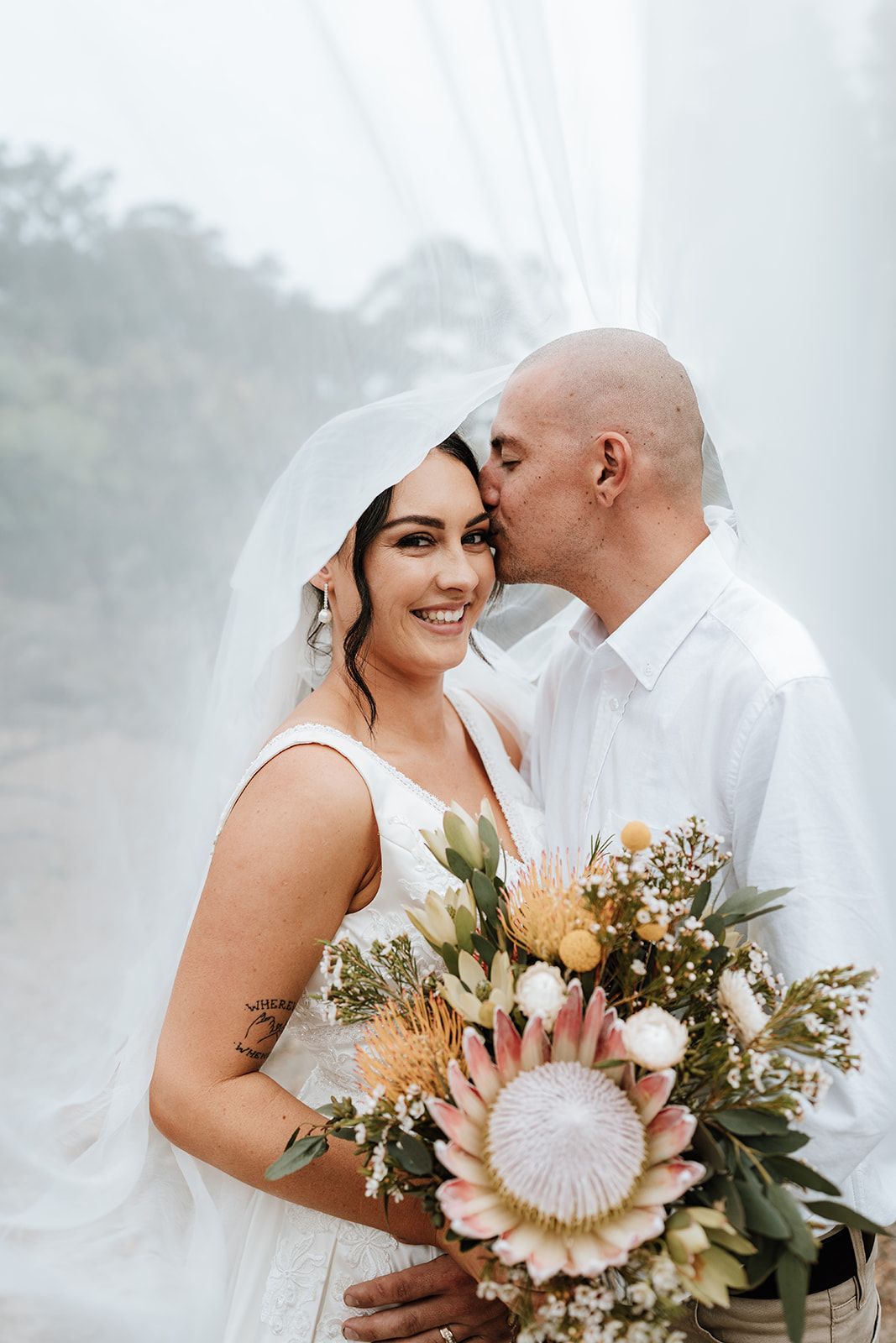 Bride and groom embracing under a veil, holding a bouquet of white and pink flowers.