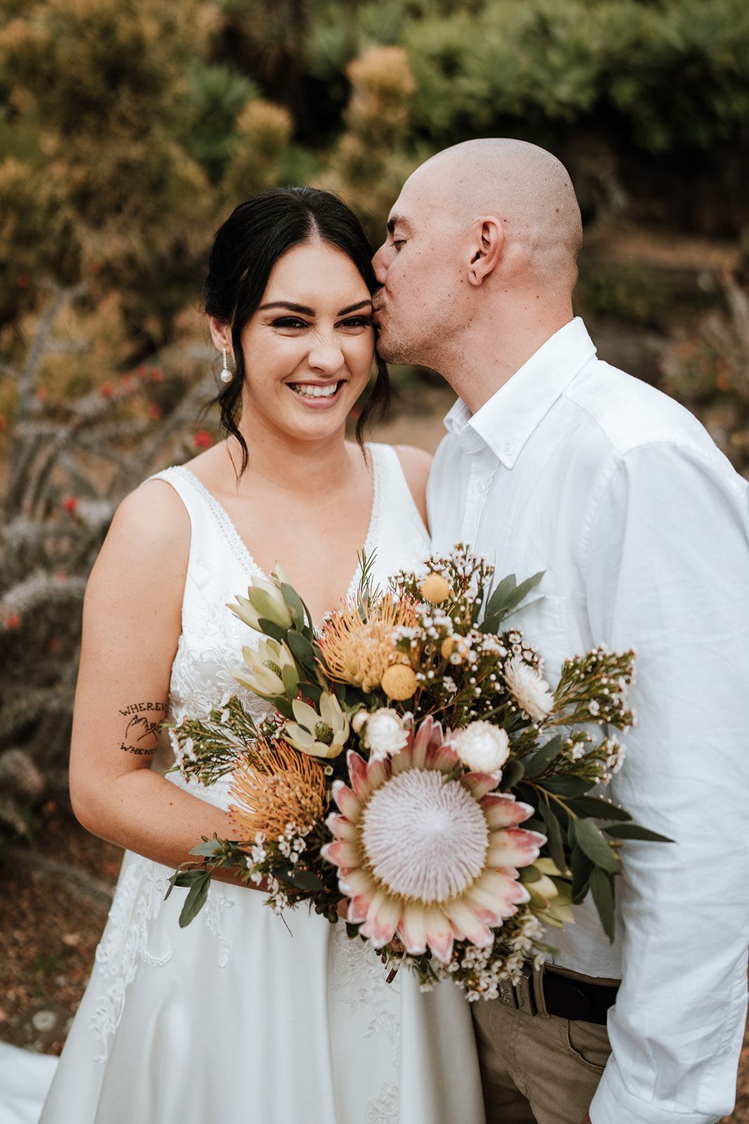 Couple in white wedding attire smiling, holding a pastel floral bouquet outdoors.