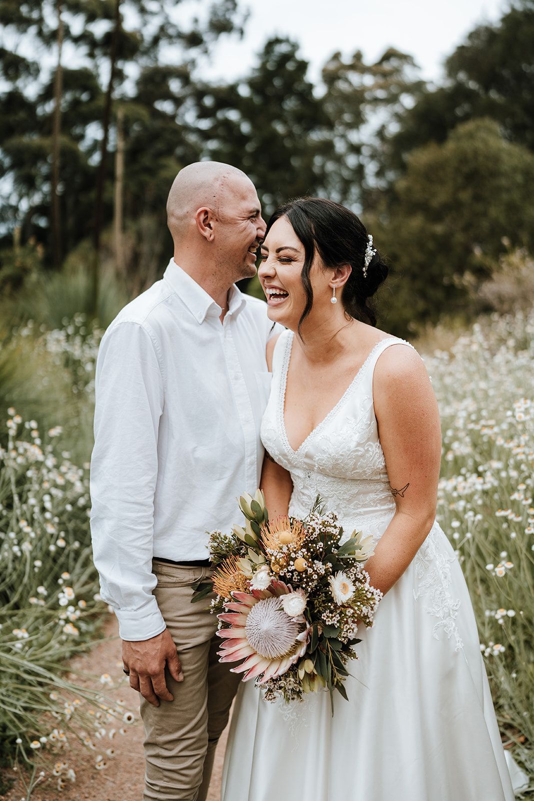 Two people in white outfits smiling outdoors, holding a bouquet in a grassy field.