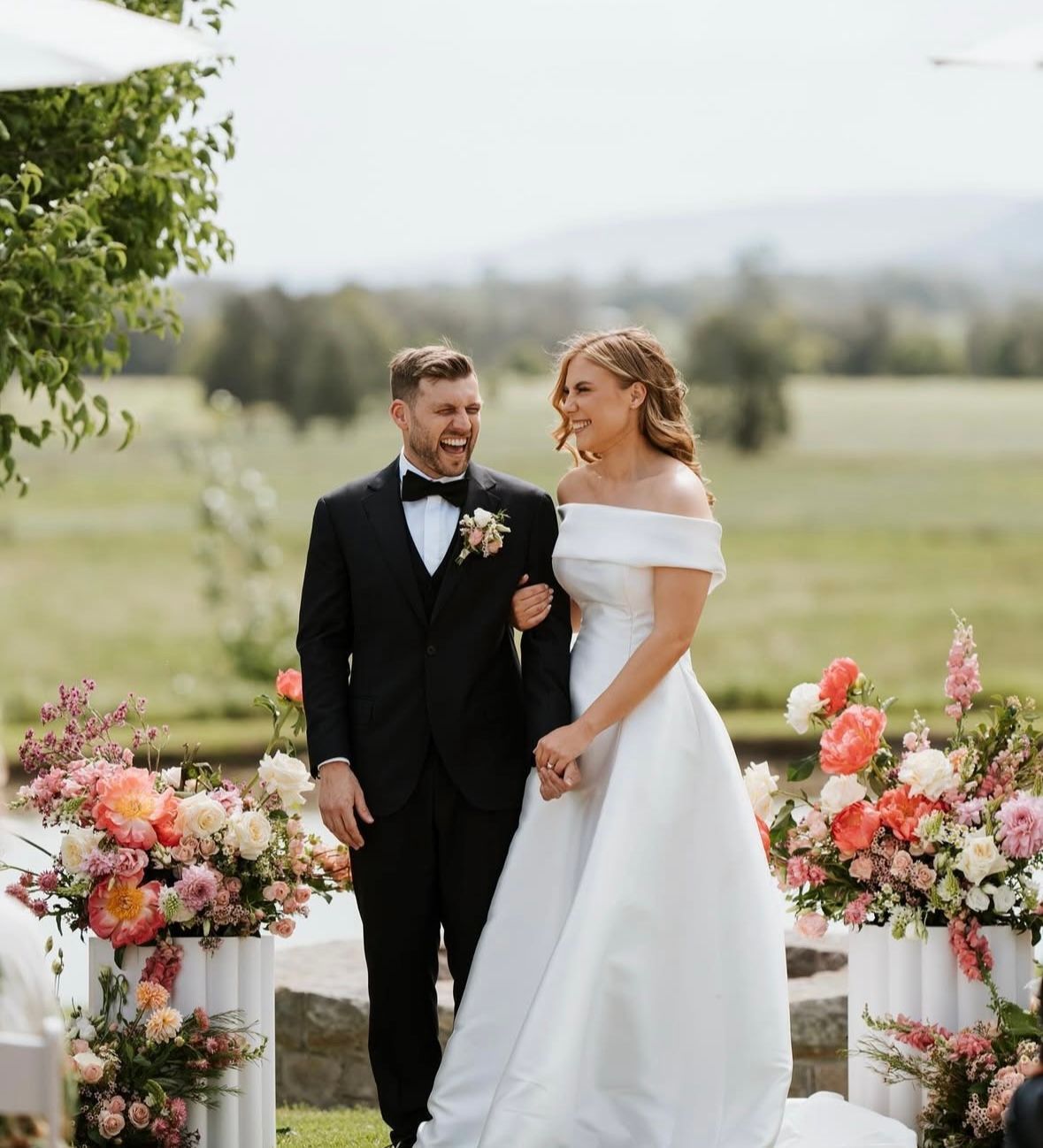 Bride and groom standing outdoors, smiling between pink floral arrangements at a wedding ceremony