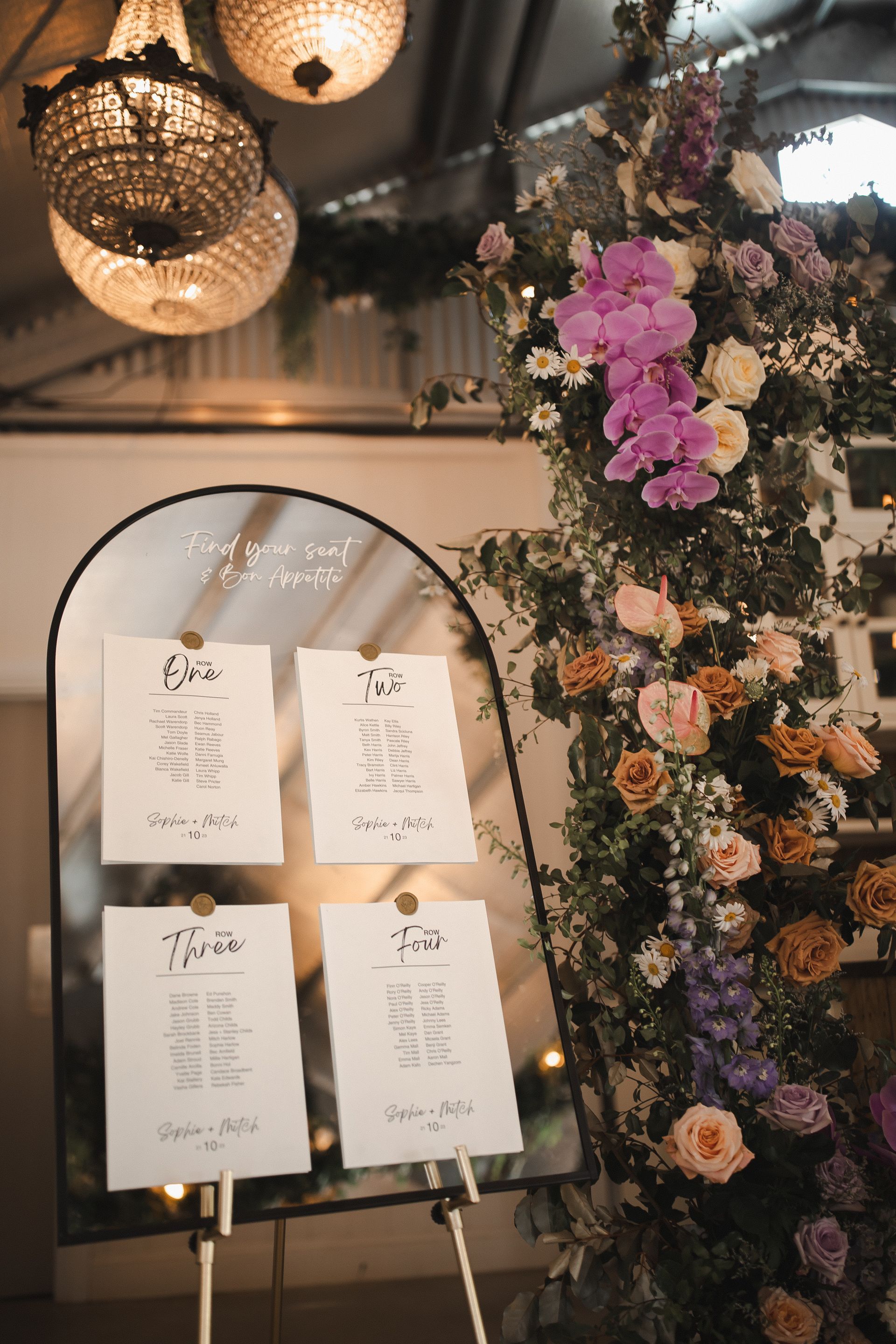 Wedding seating chart on an easel beside a floral arch with purple and peach flowers.