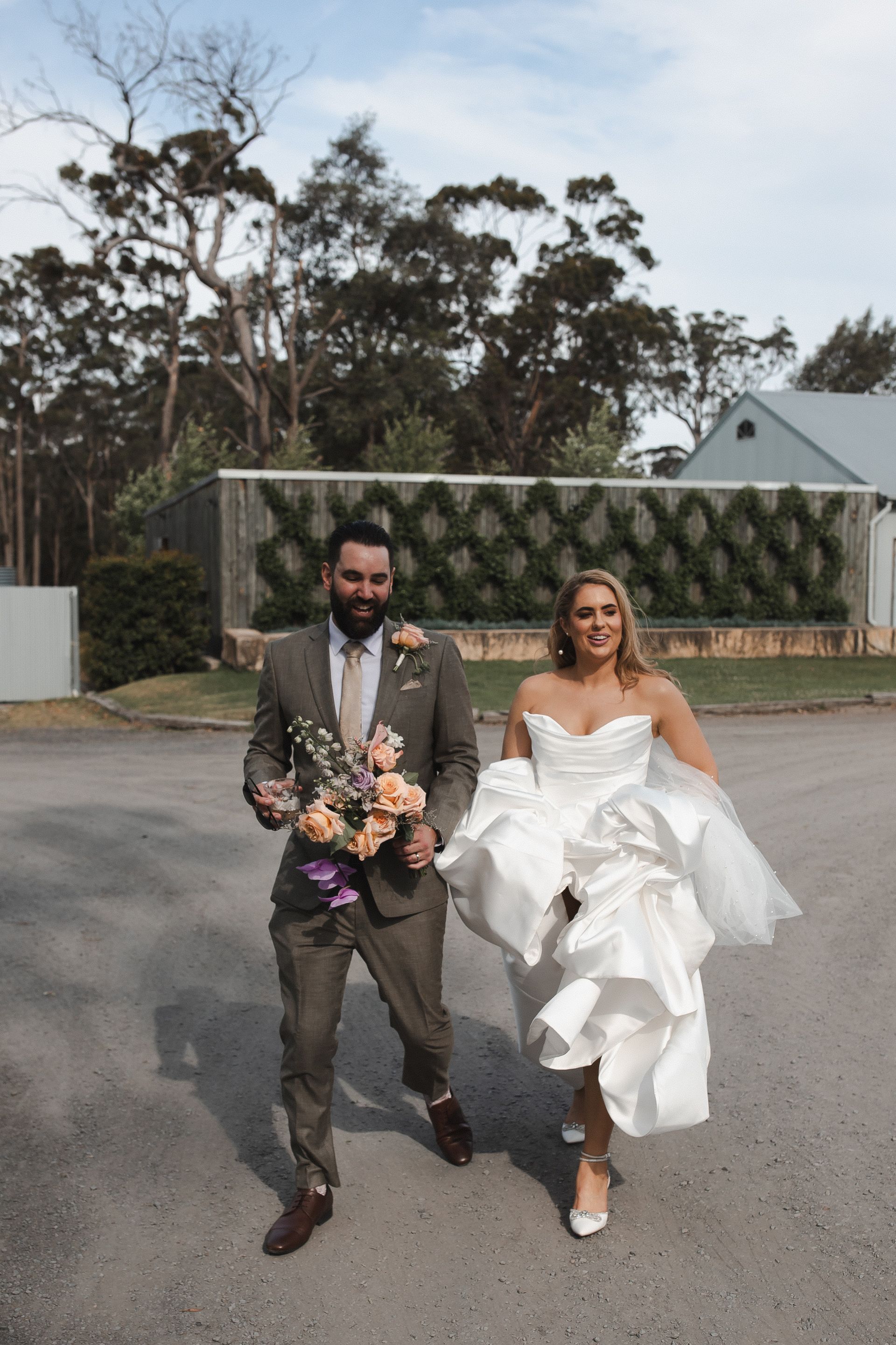 Bride and groom walking outdoors in wedding attire, smiling and holding bouquets
