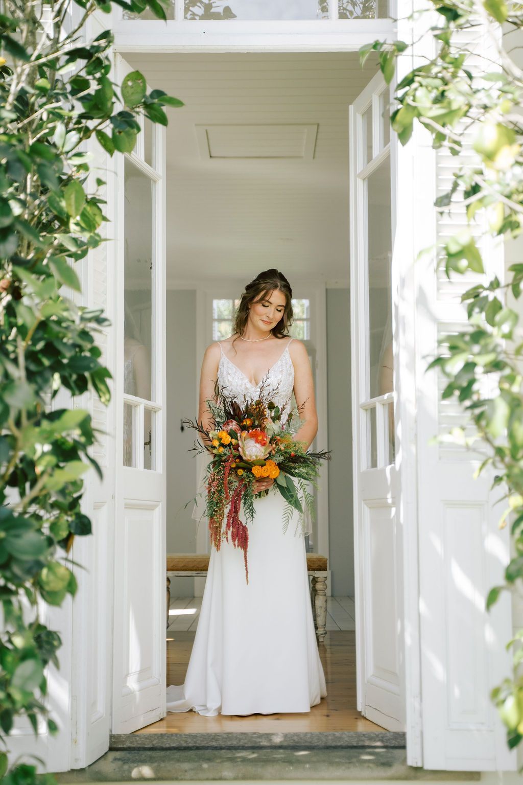 Bride in a white dress standing in a doorway holding a colorful bouquet, framed by greenery