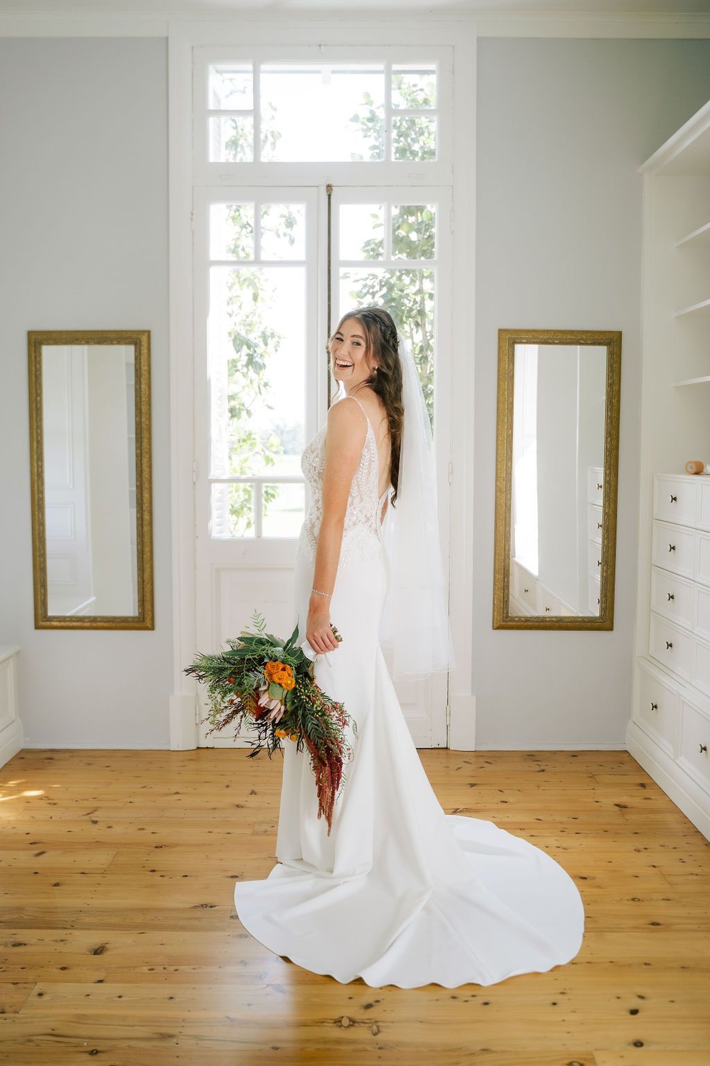 Bride in a white gown holding a colorful bouquet in a bright room with tall windows