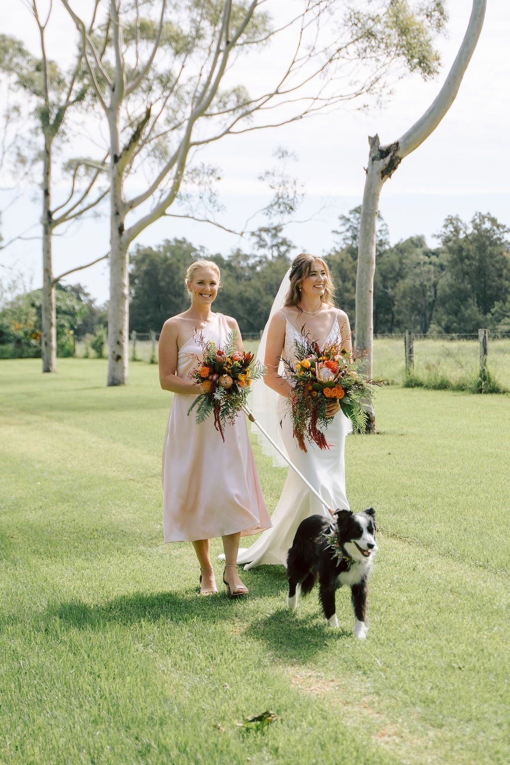 Two women in pastel dresses walk on a golf course, carrying bouquets, with a black-and-white dog ahead.