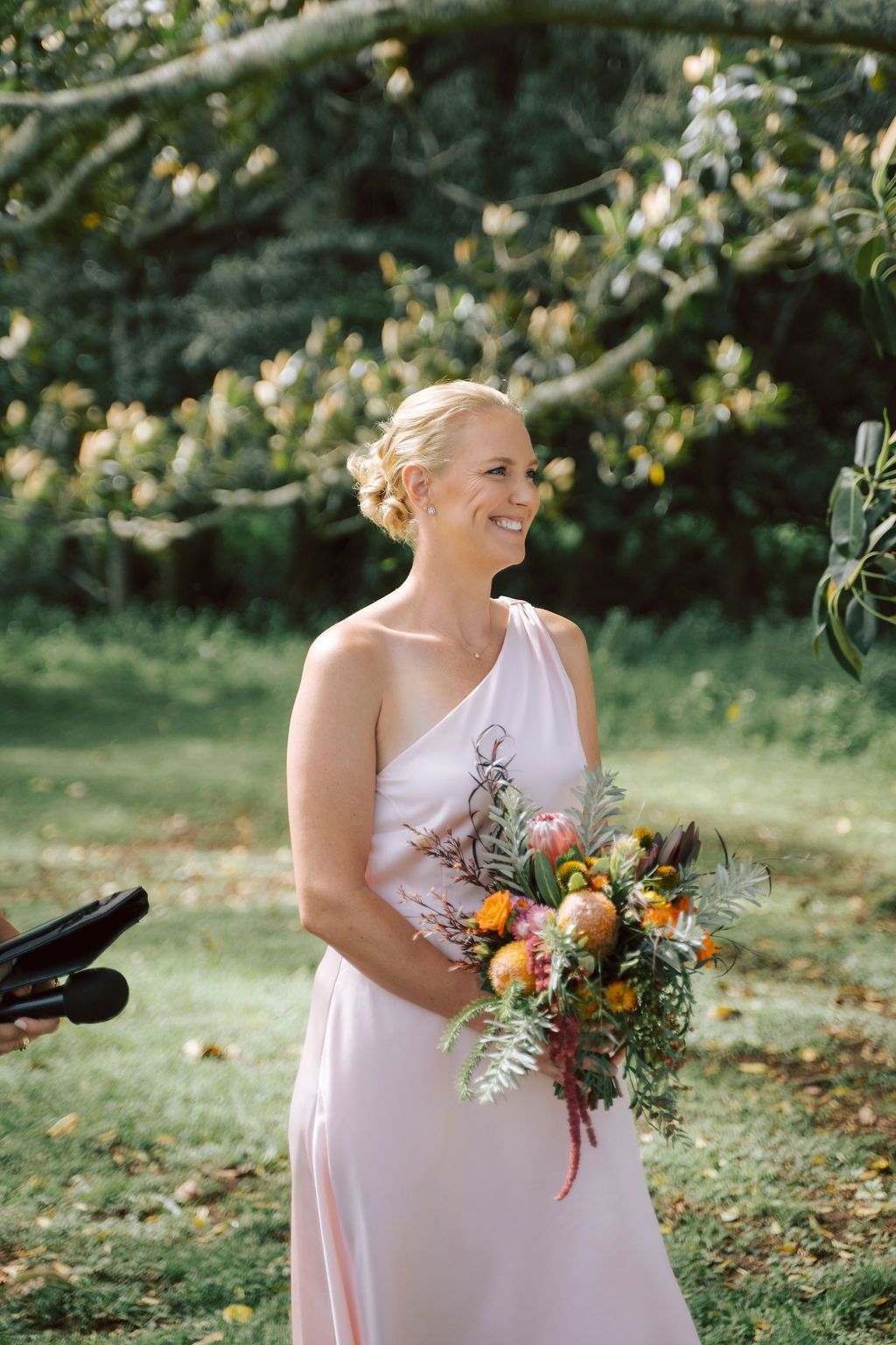 Person in a blush dress holding a colorful bouquet outdoors under trees