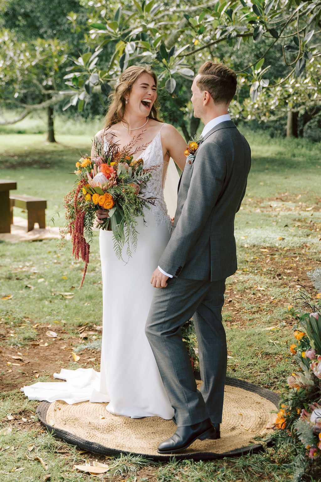 Bride and groom laughing in a garden wedding, standing on a rug beneath trees with a bouquet and boutonniere.