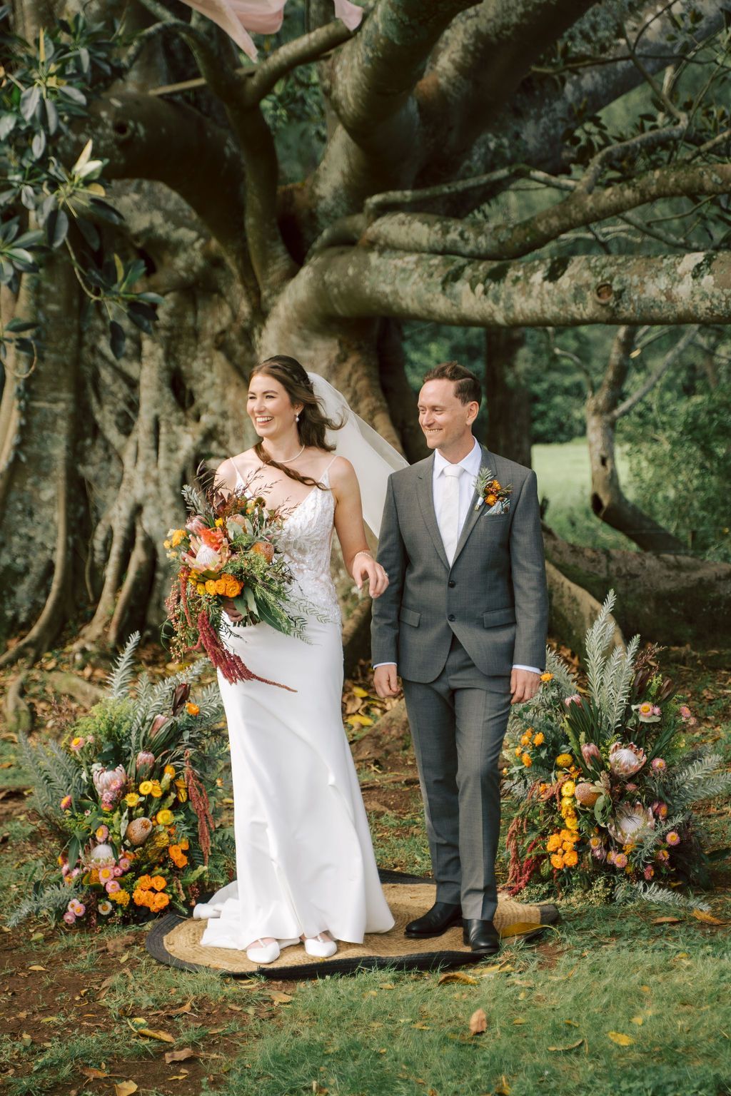 Bride and groom standing under a large tree, surrounded by colorful floral arrangements outdoors.