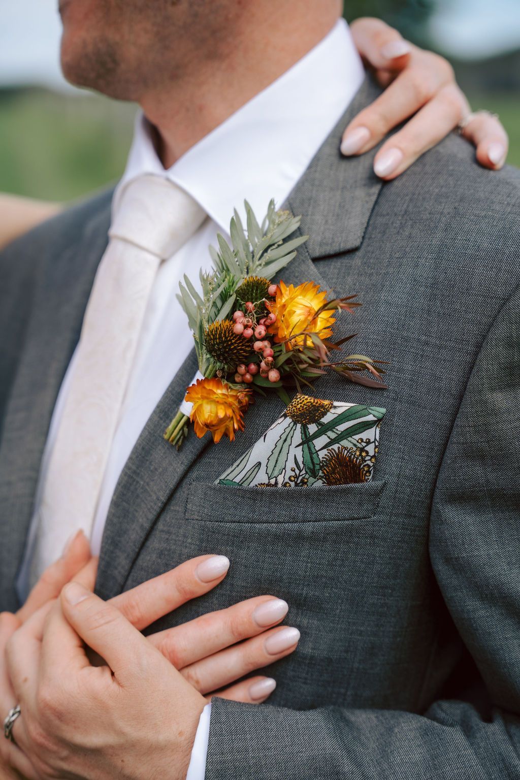 Close-up of a groom in a gray suit with a white tie and orange boutonniere, hands gently clasped.