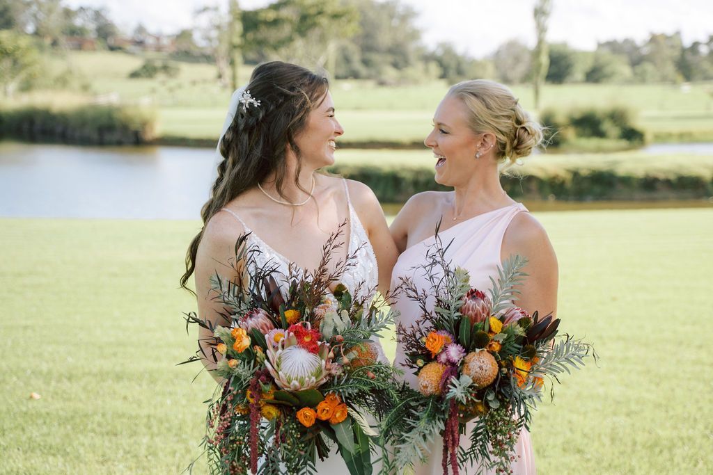 Two brides in white dresses holding colorful bouquets, smiling by a lakeside
