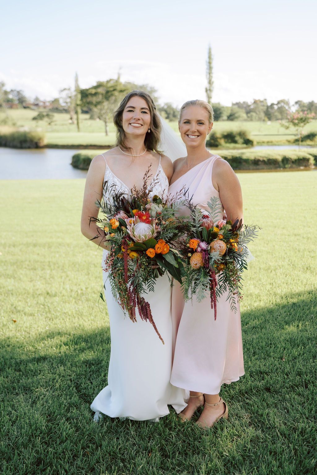 Two people in white dresses holding colorful bouquets, standing on a grassy lawn outdoors.