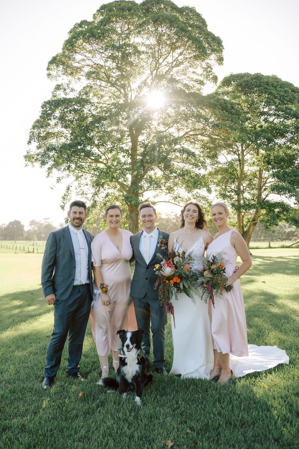 Five people in formalwear stand on a grassy lawn with bouquets, backlit by a large tree at sunset.