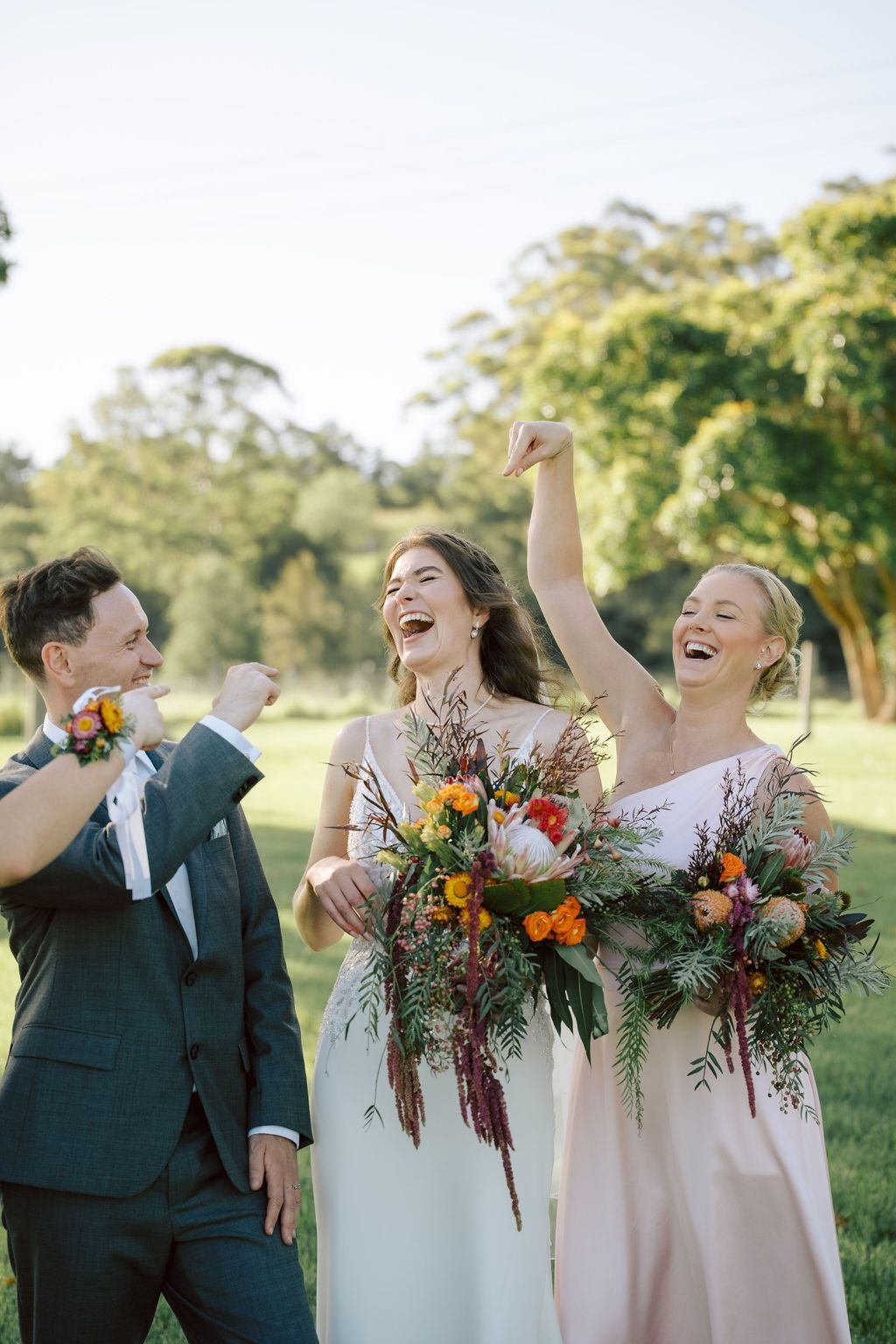 Three people in formalwear celebrating outdoors, holding colorful bouquets and smiling.