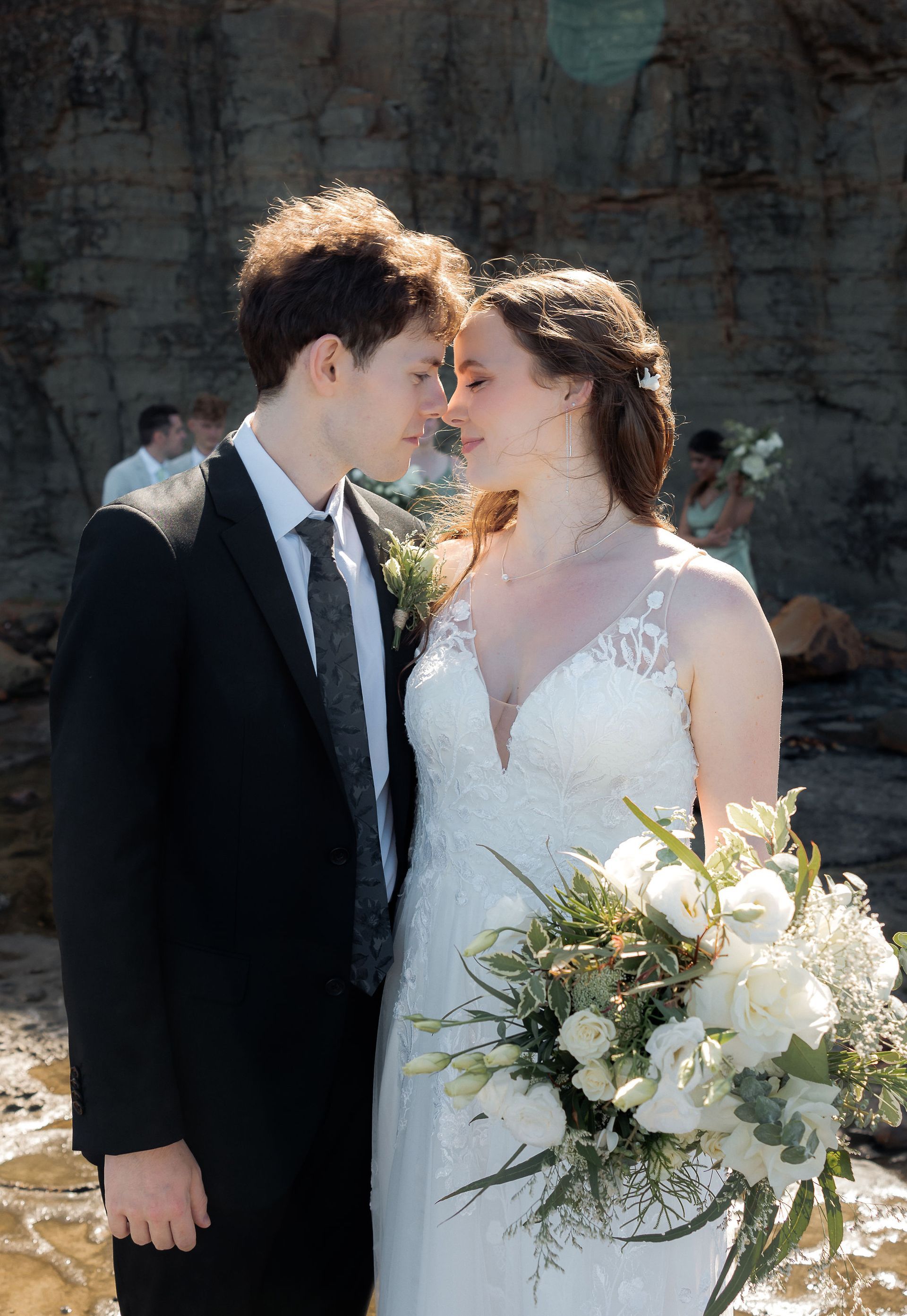 Wedding couple in white dress and dark suit, touching foreheads outdoors with bouquet in hand