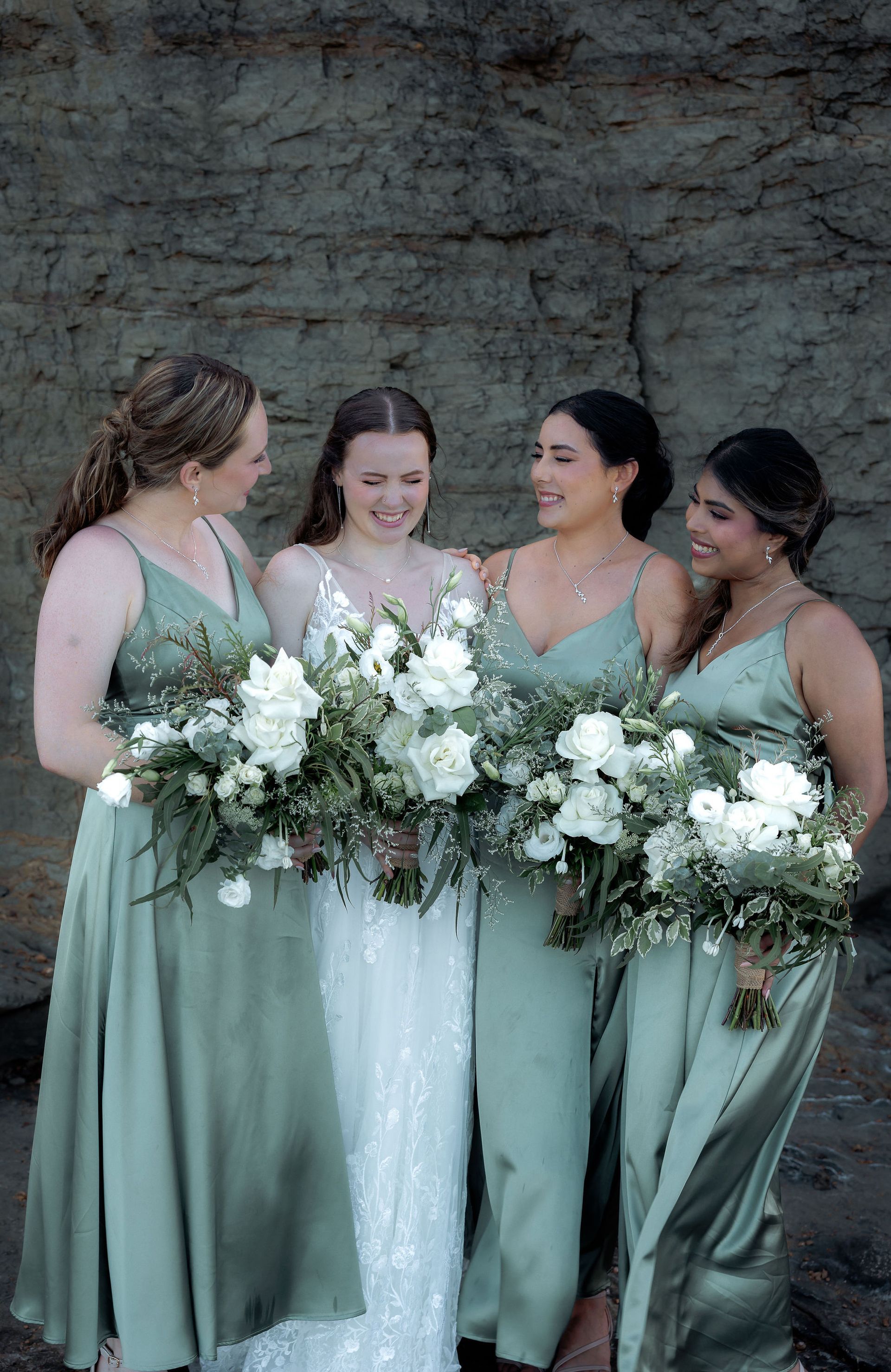 Four women in sage dresses holding white flower bouquets against a stone wall