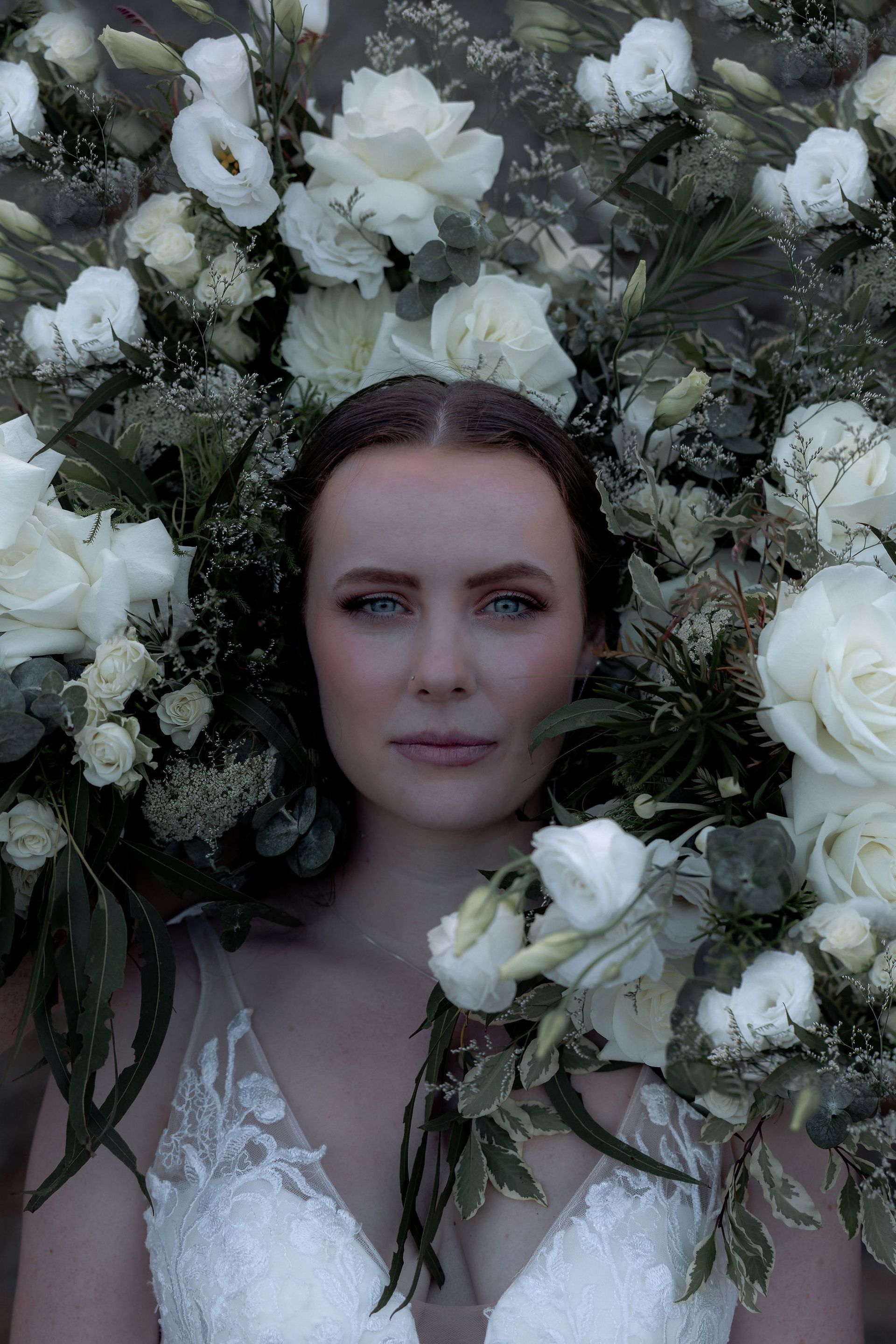 Woman framed by white roses and greenery, with a calm expression and dark hair, against a floral backdrop.