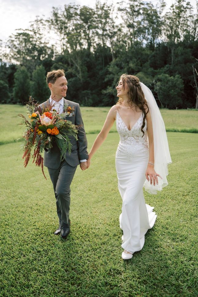 Two brides in wedding attire walking hand in hand across a grassy field with a bouquet