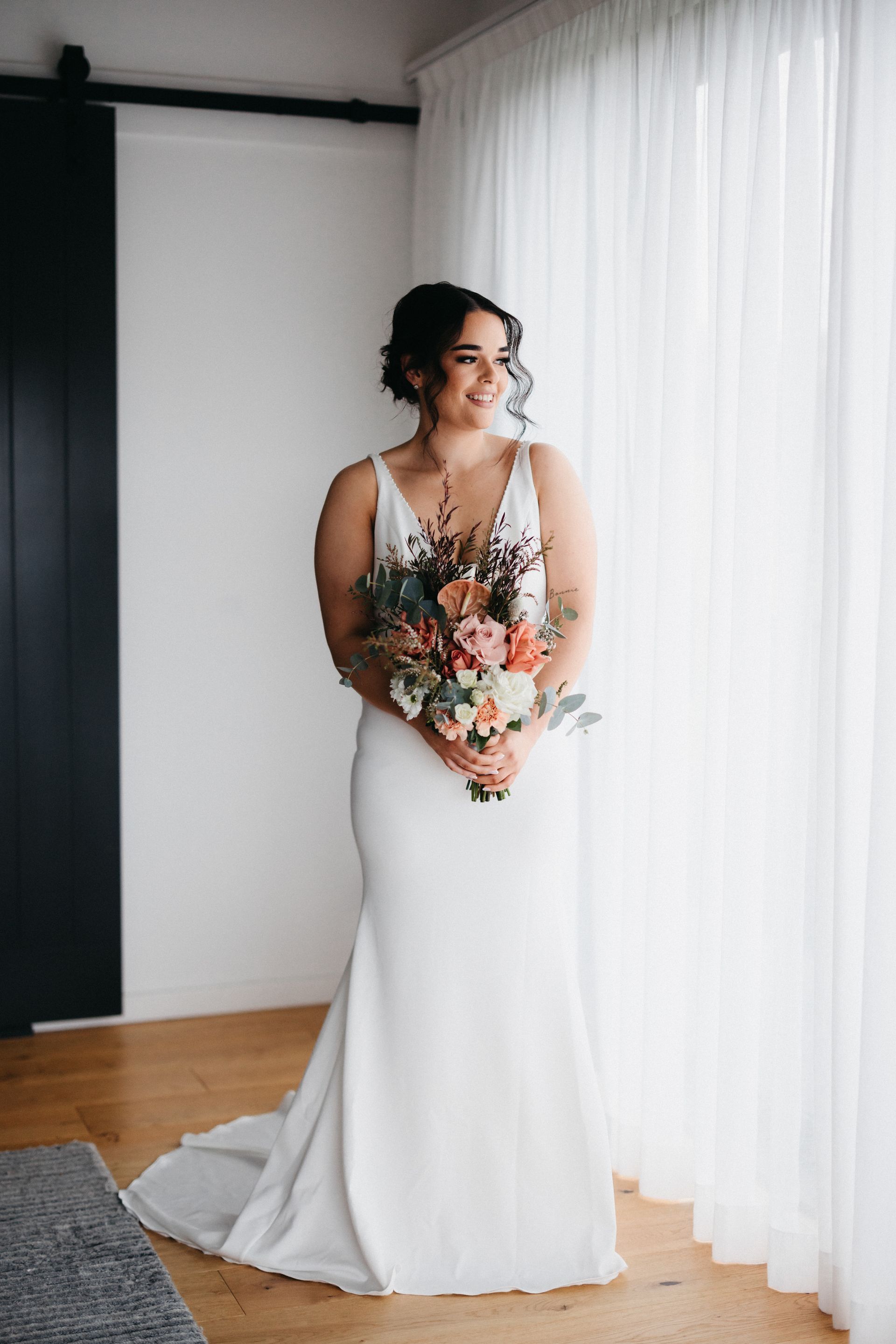 Bride in a white gown holding a bouquet, standing by a bright curtained window.