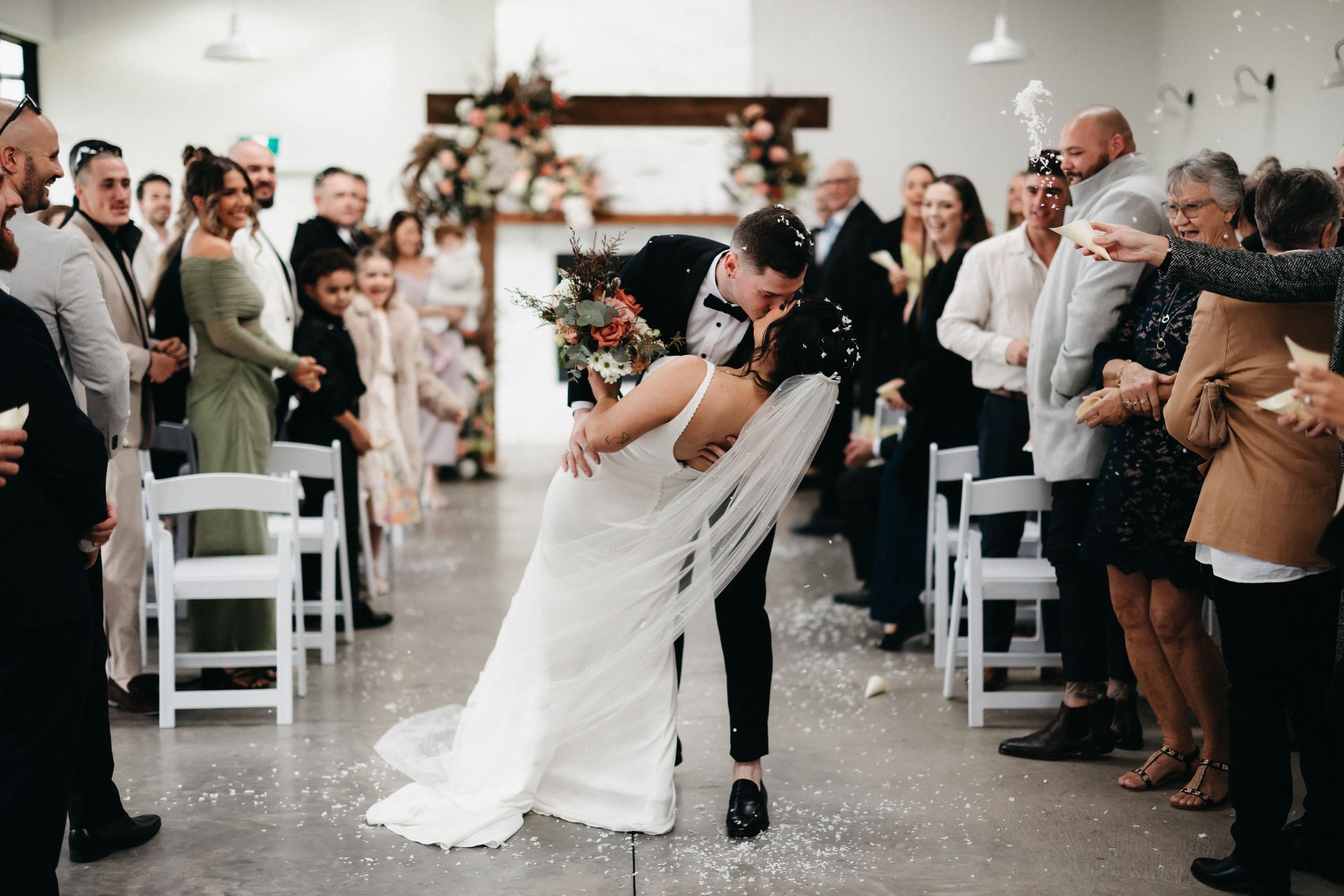 Bride and groom kiss in a decorated wedding aisle as guests cheer and clap nearby.