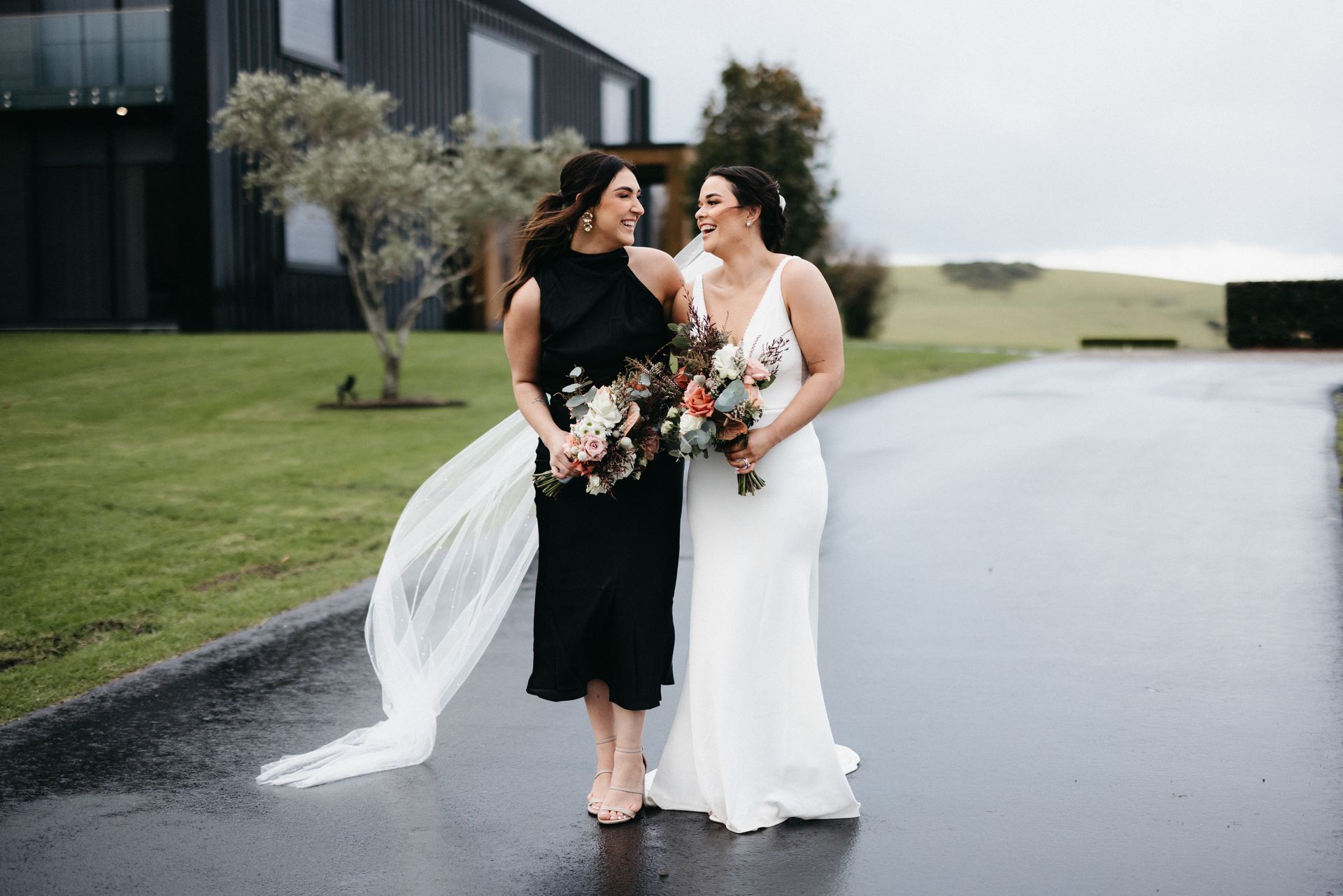 Two brides in black and white dresses walking with bouquets on a rainy road