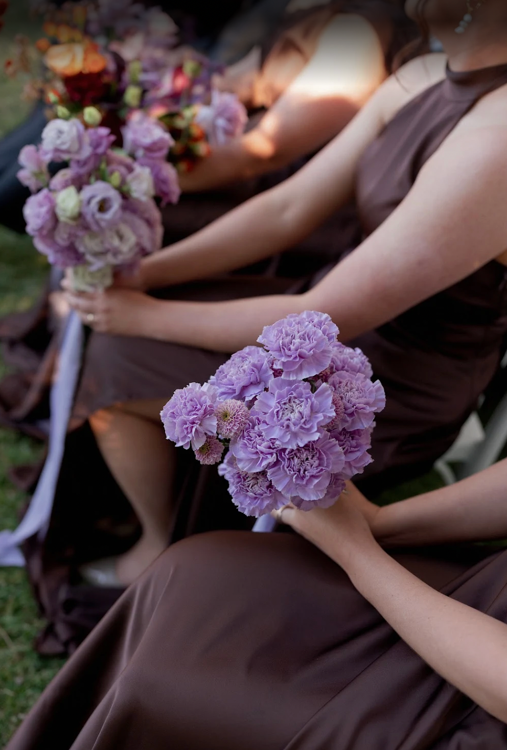 People in brown dresses holding purple flower bouquets in a soft, blurred setting