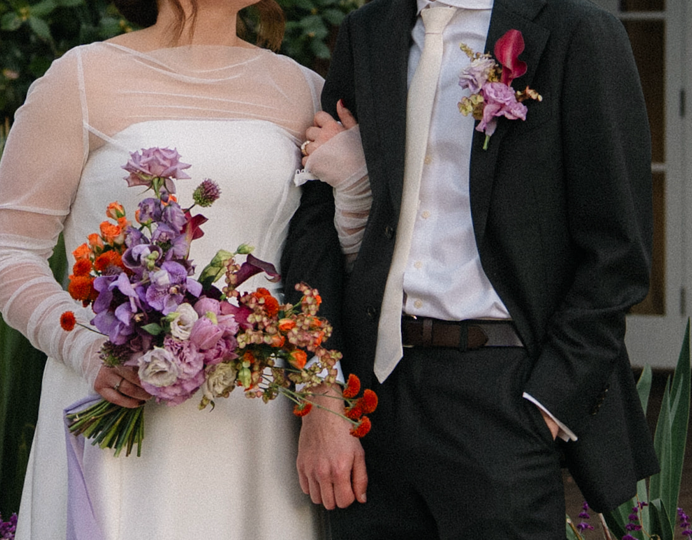 Bride and groom holding hands, wearing white and dark suits, with colorful bouquets and corsages.