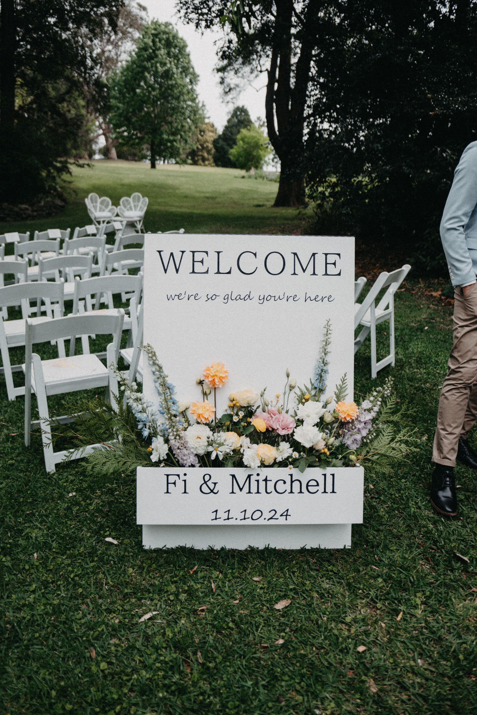 Welcome sign with flowers for “Fi & Mitchell,” displayed outdoors on a lawn with white chairs.