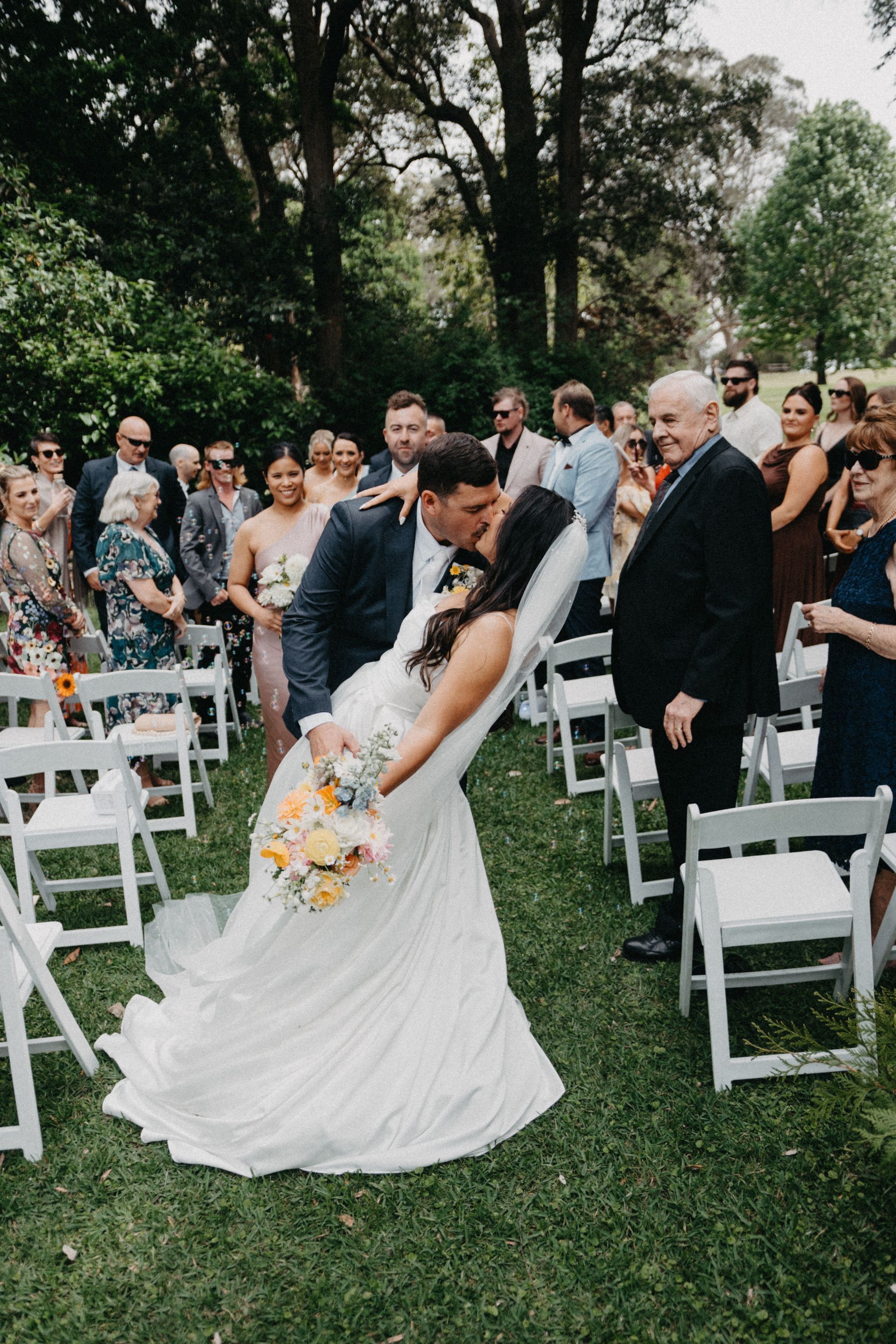 Bride in a white gown and groom in a dark suit exchange vows outdoors at a garden wedding.