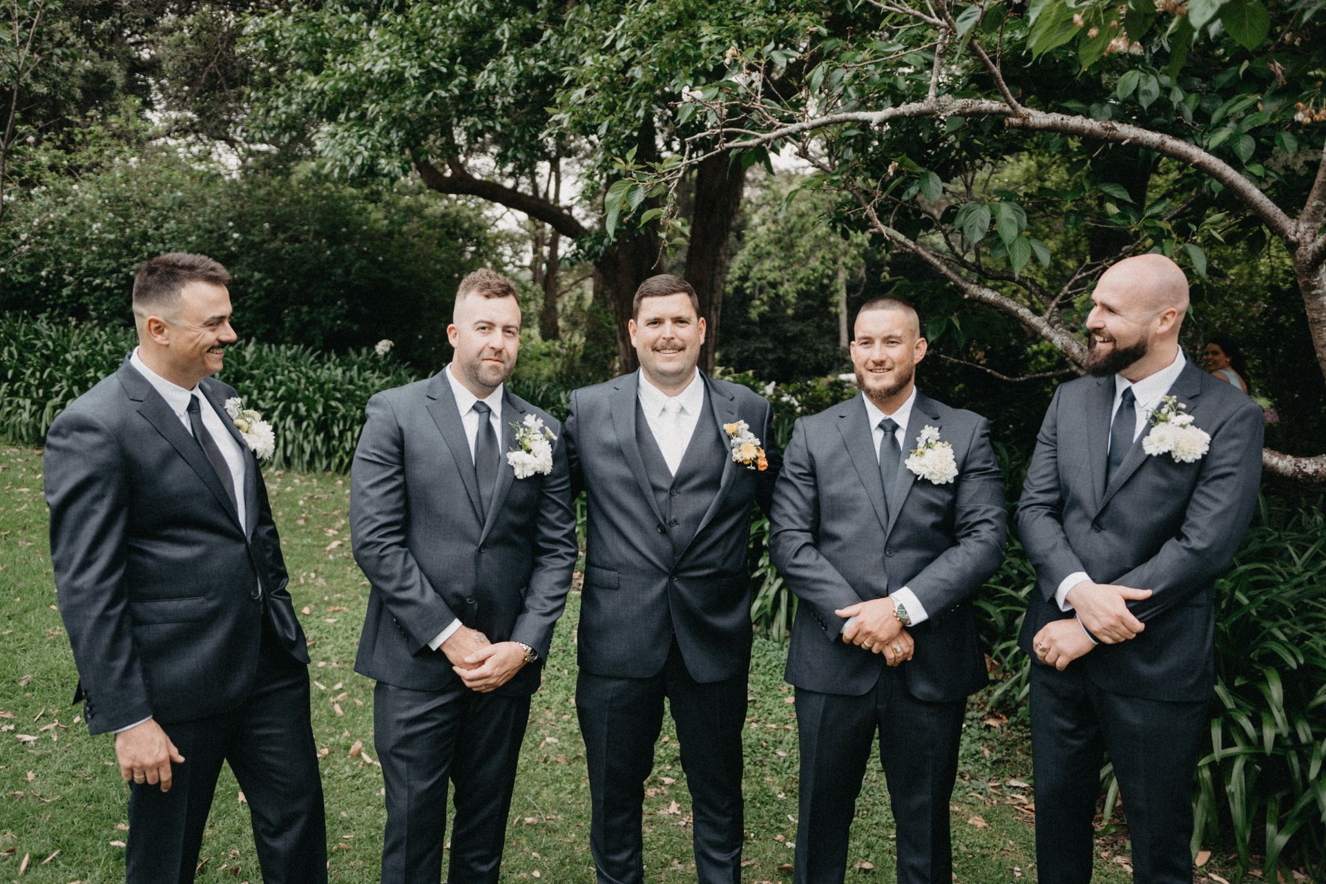 Five men in dark suits stand together outdoors in front of trees, smiling at a formal event.