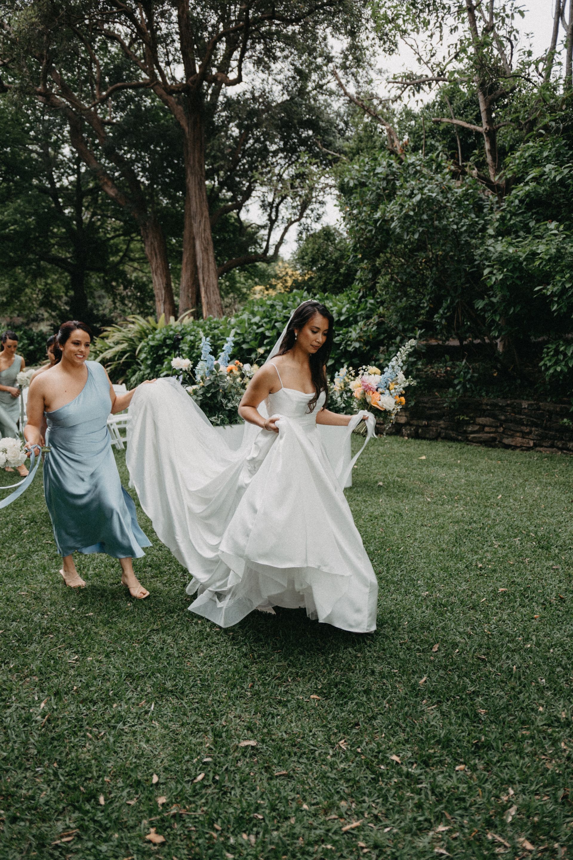Bride in a white gown runs across a grassy garden with a bouquet, followed by a bridesmaid in a light blue dress.