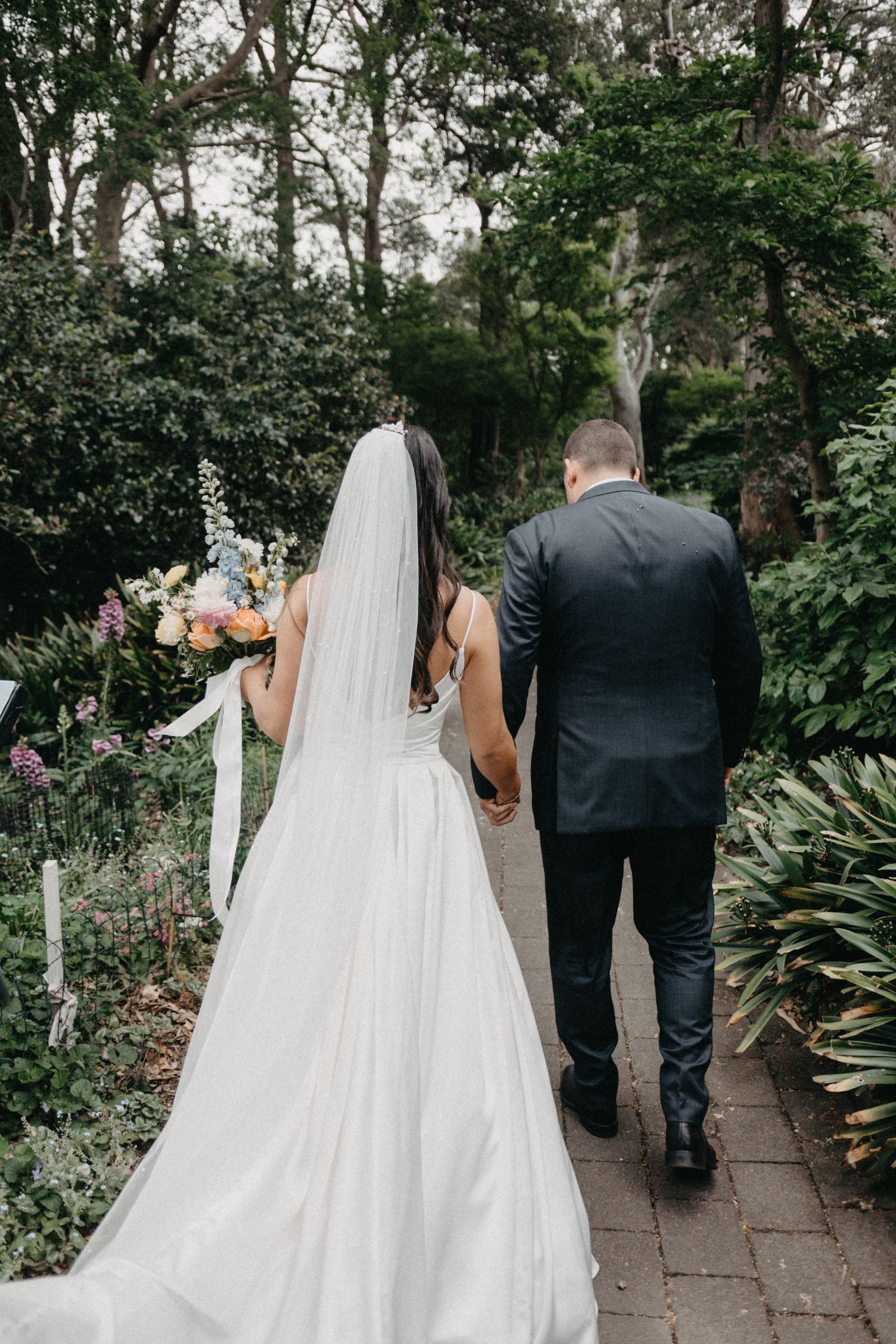 Bride in a white gown and veil walking with groom in a dark suit along a garden path, back view