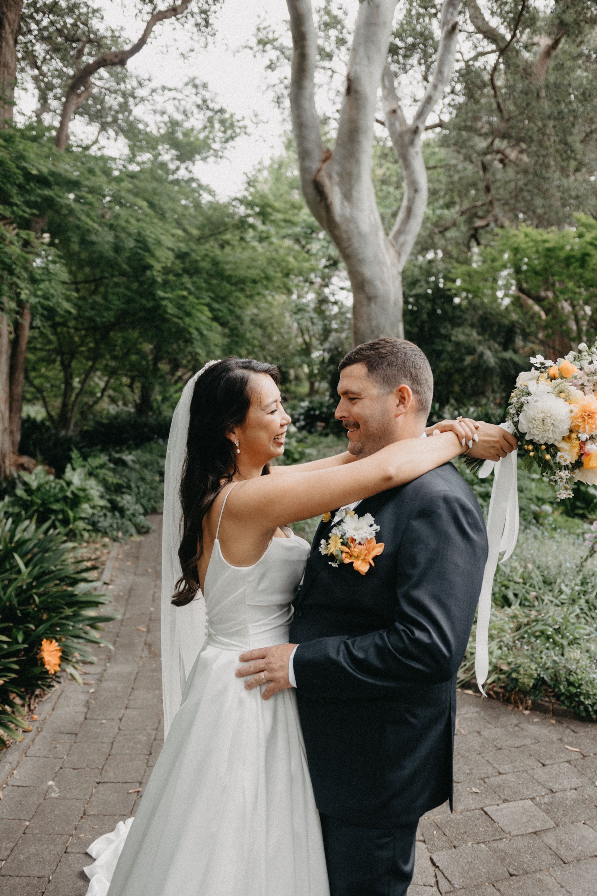 Couple in wedding attire embracing outdoors on a garden path, holding a bouquet.
