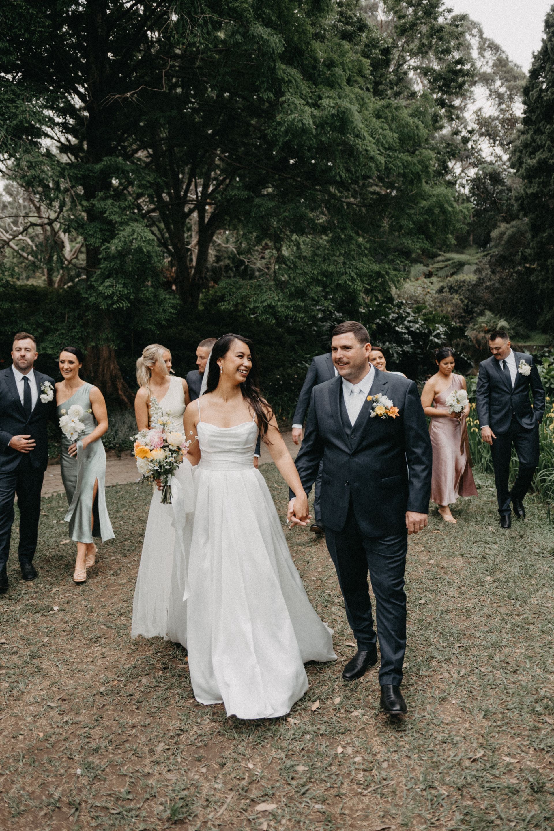Wedding party walking outdoors along a wooded path, bride in white dress and groom in dark suit leading the group