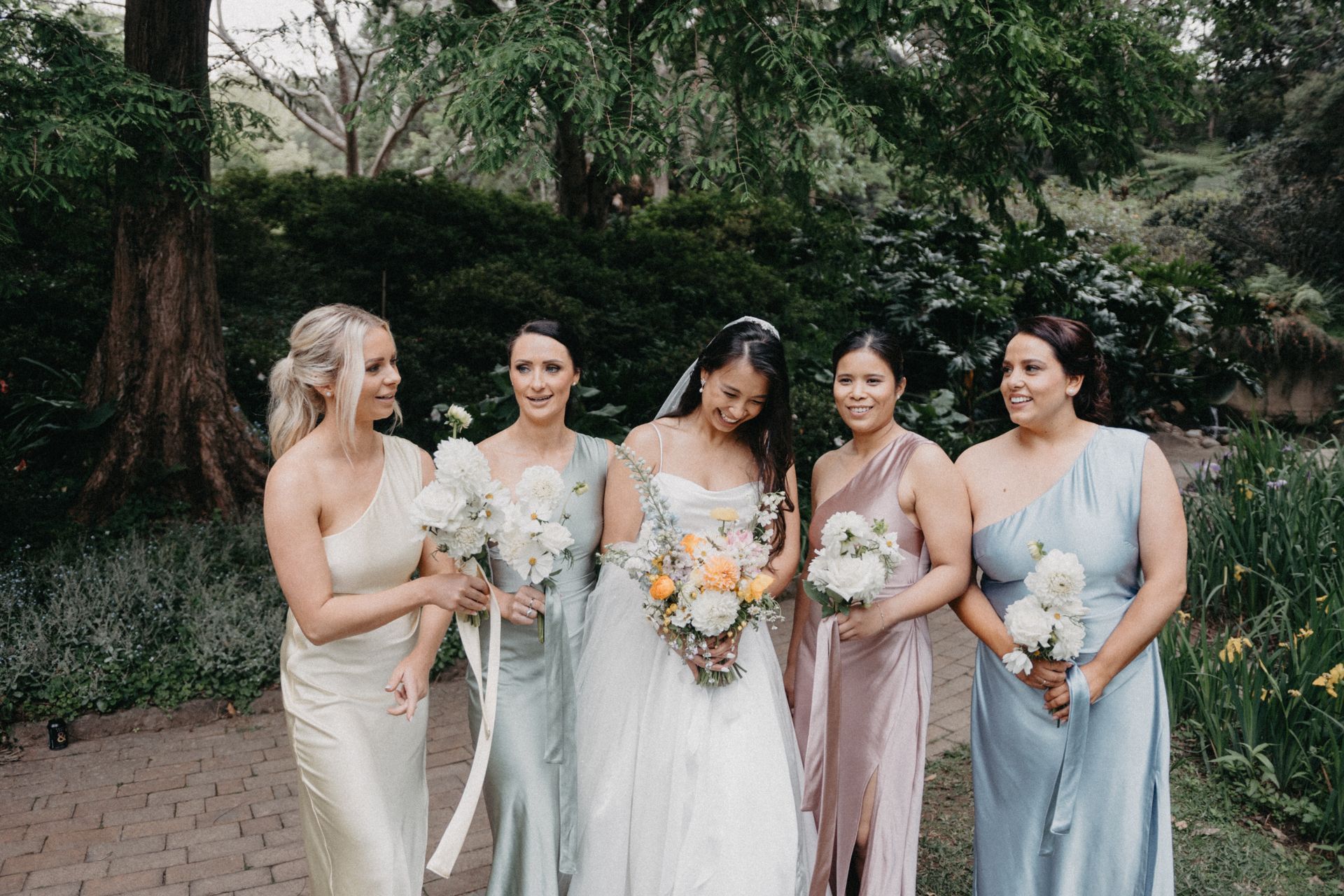 Five women in pastel dresses hold bouquets outdoors at a wedding, smiling in a garden.